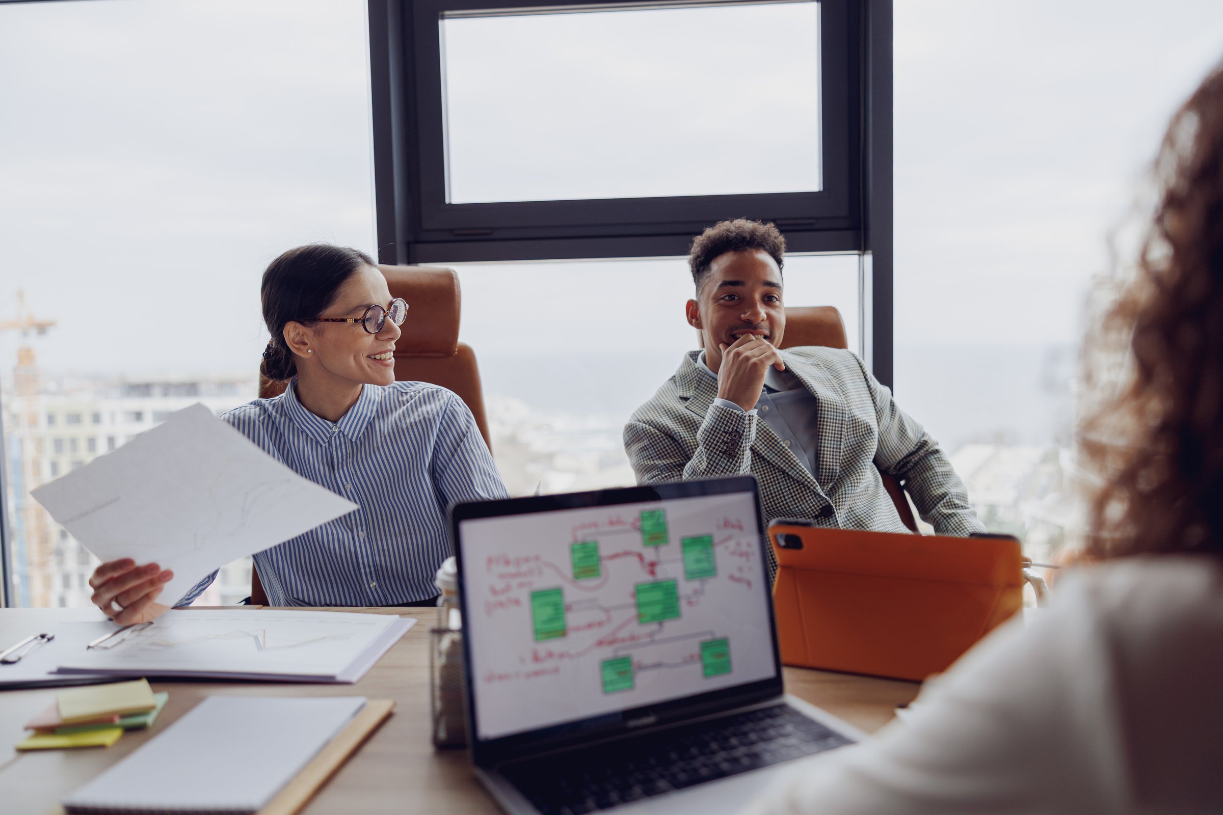 A diverse business team discusses thoughtfully, using modern technology with a city skyline view behind them