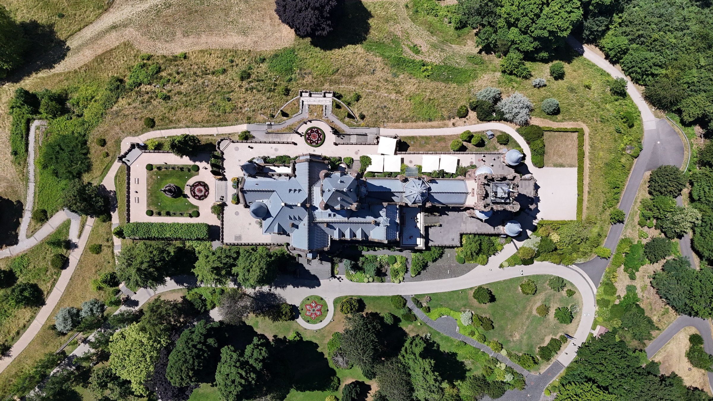 Aerial view of a historic mansion surrounded by landscaped gardens and pathways on a sunny day, showcasing architectural elegance and natural beauty.