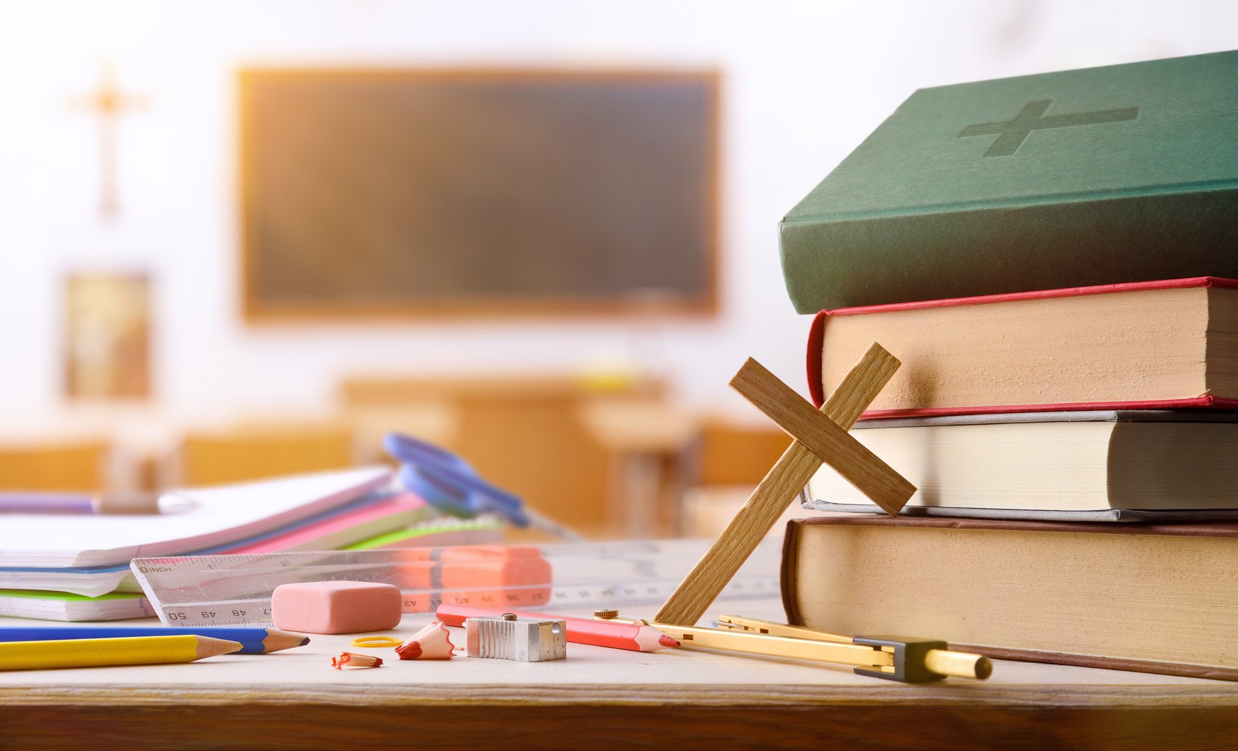 Background with tools and religious educational materials such as notebooks, pencils, books, and Bible on a wooden table with a classroom in the background with desks and blackboard. Front view.