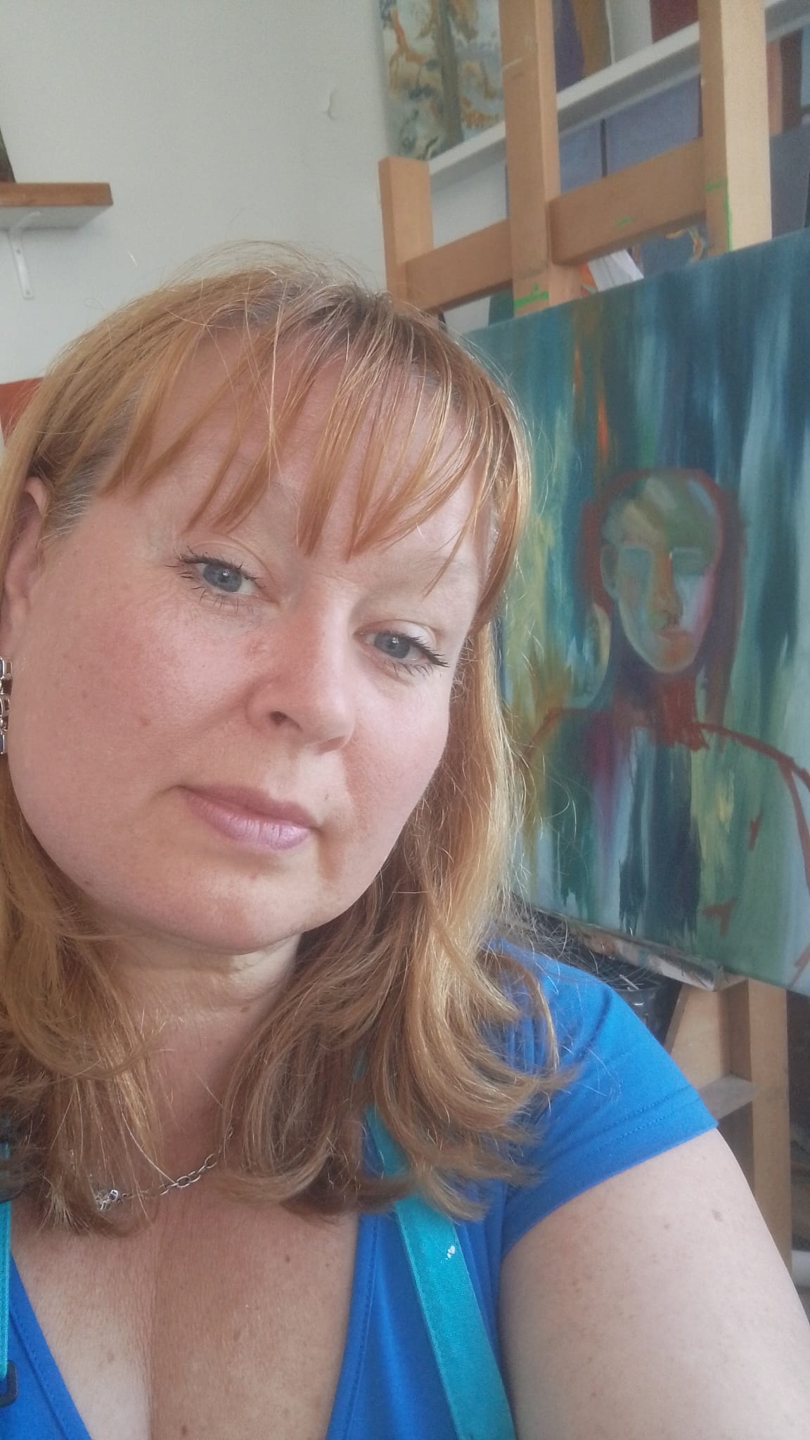 Woman with auburn hair takes a selfie in front of an abstract painting on an easel in an art studio.