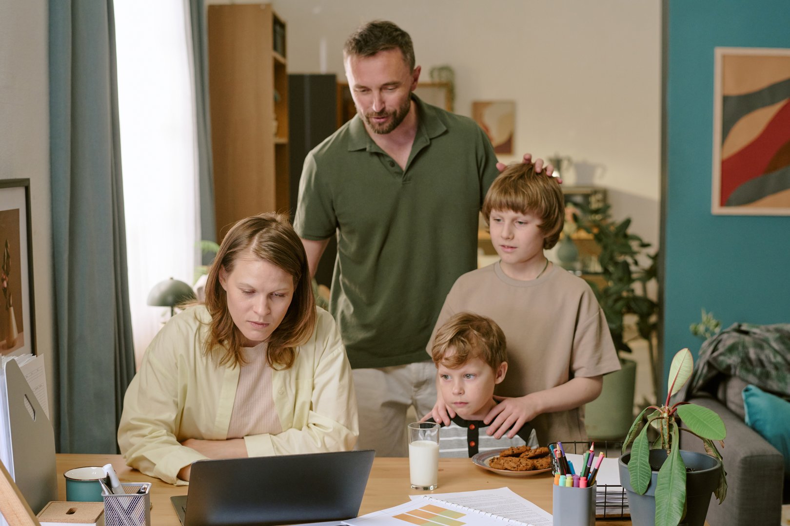 Caucasian middle aged woman working on laptop at home office desk while Caucasian middle aged man and two Caucasian boys standing nearby, one boy holding glass of milk and plate of cookies