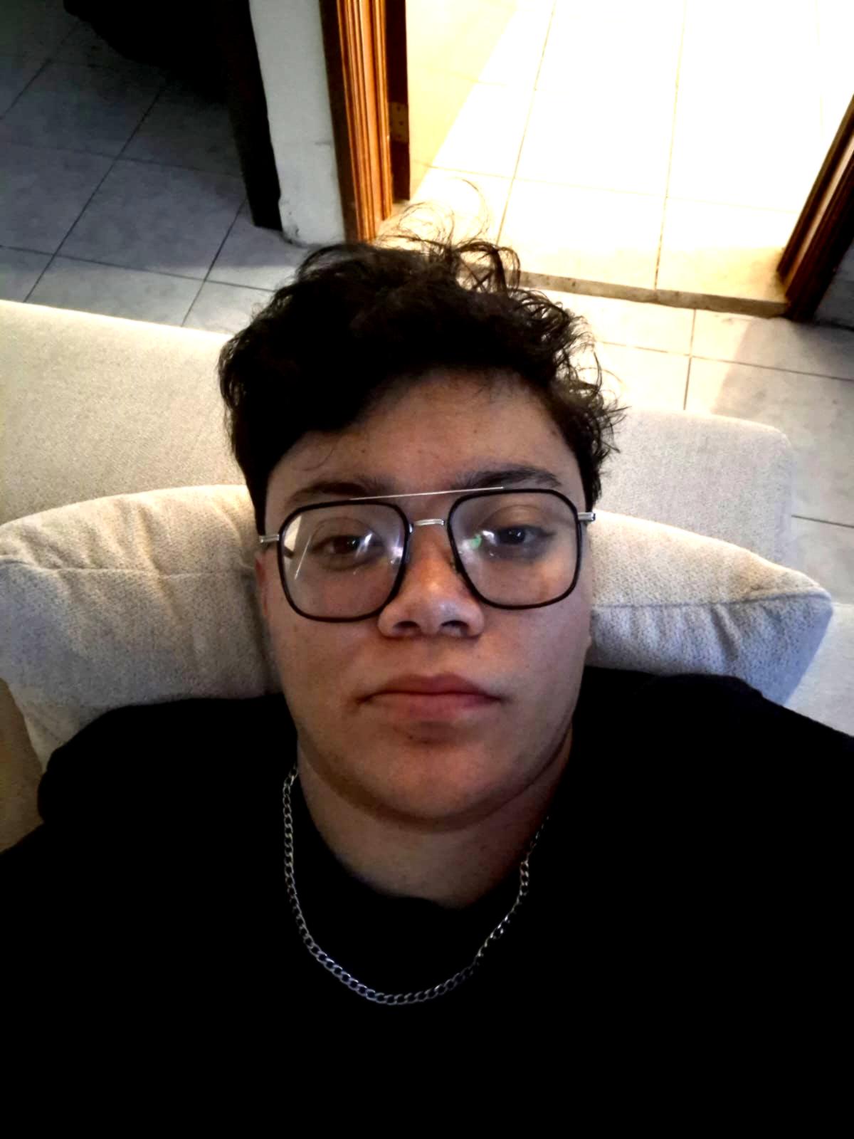 Person with short curly hair and glasses lying on a cushion indoors, wearing a dark top and a chain necklace.