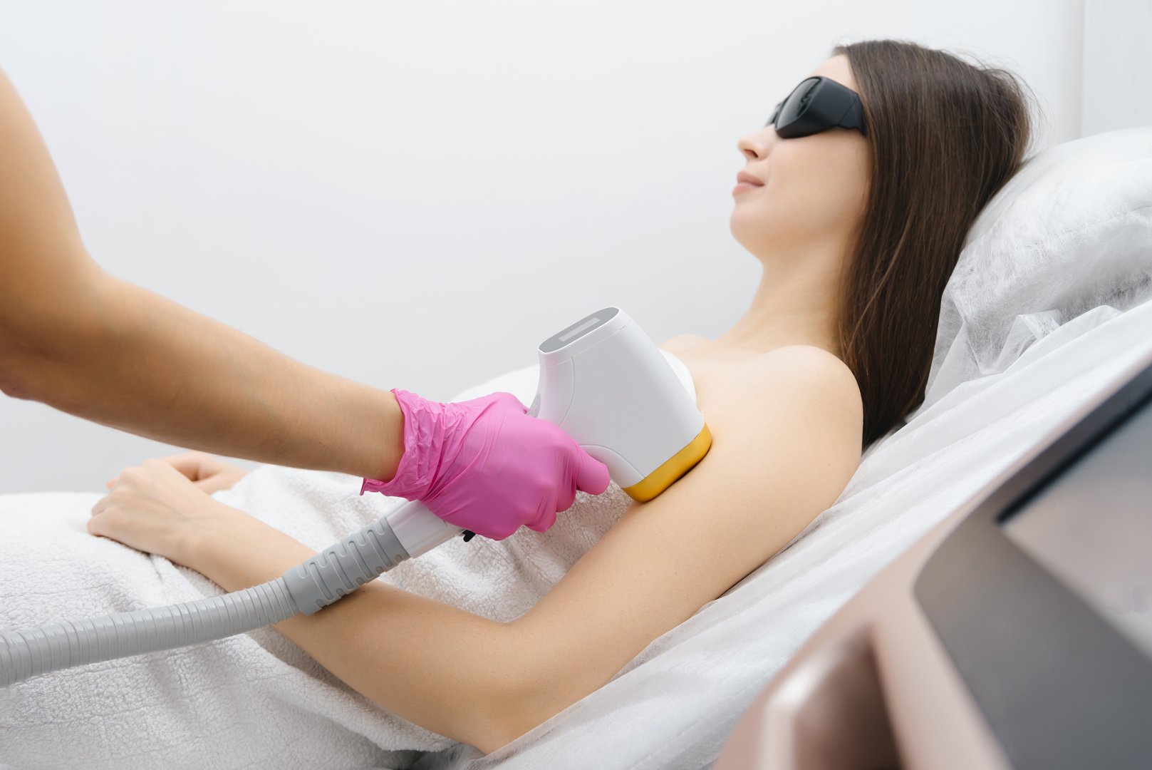 laser epilation, hair removal procedure, young beautiful caucasian smiling brunette woman lying on couch in beauty salon, arm epilation, pink gloves, black glasses, spa and cosmetology concept