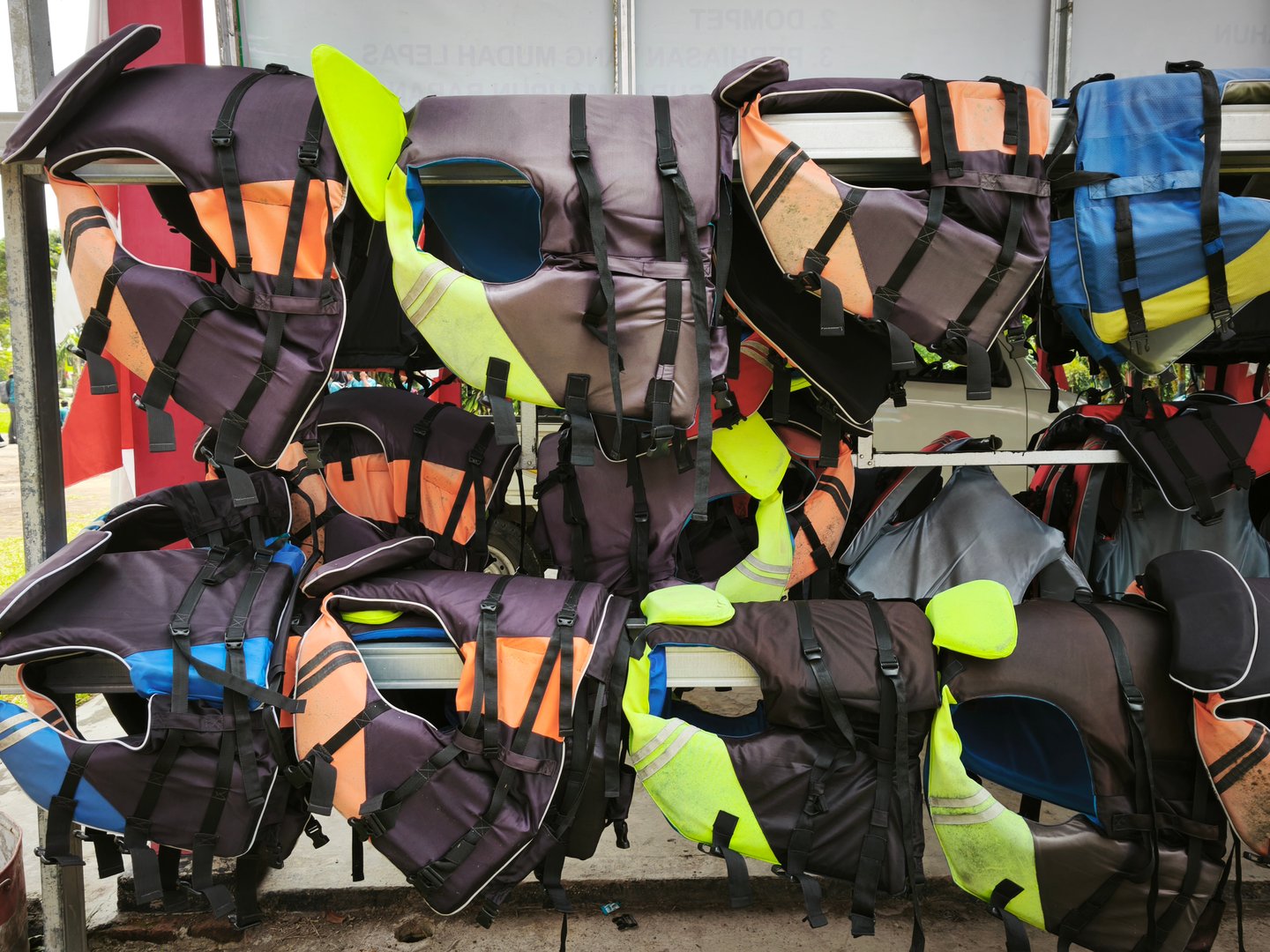 Stack of Colorful Personal Flotation Devices (PFDs) Hanging on a Rack: A Messy Array of Life Vests in Dark Tones with Bright Orange, Neon Yellow, and Teal Accents, Ready for Rental or Use in Water Sports and Boating Safety Activities