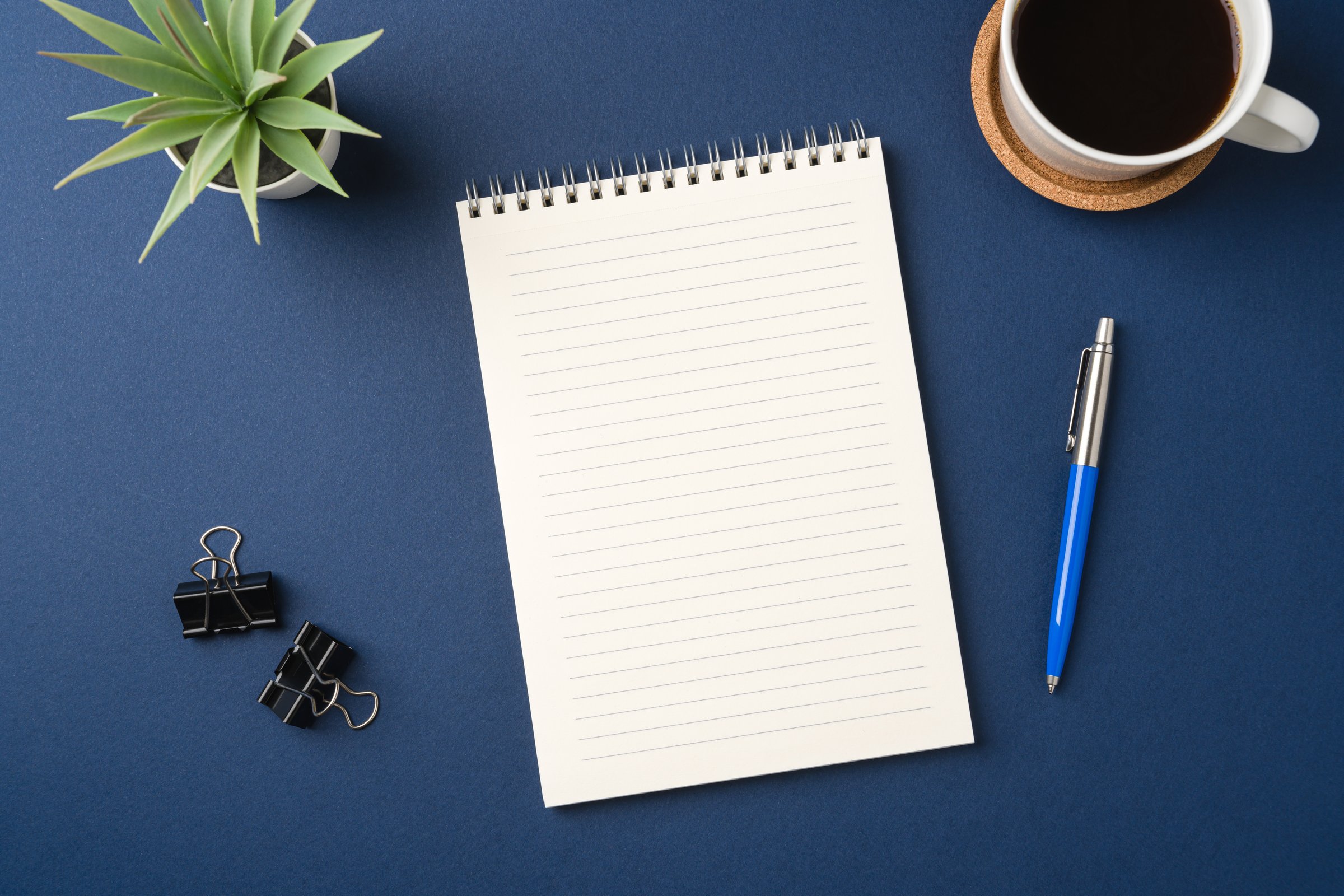 Top view of a workspace with a blank notebook, pen, coffee, and plant