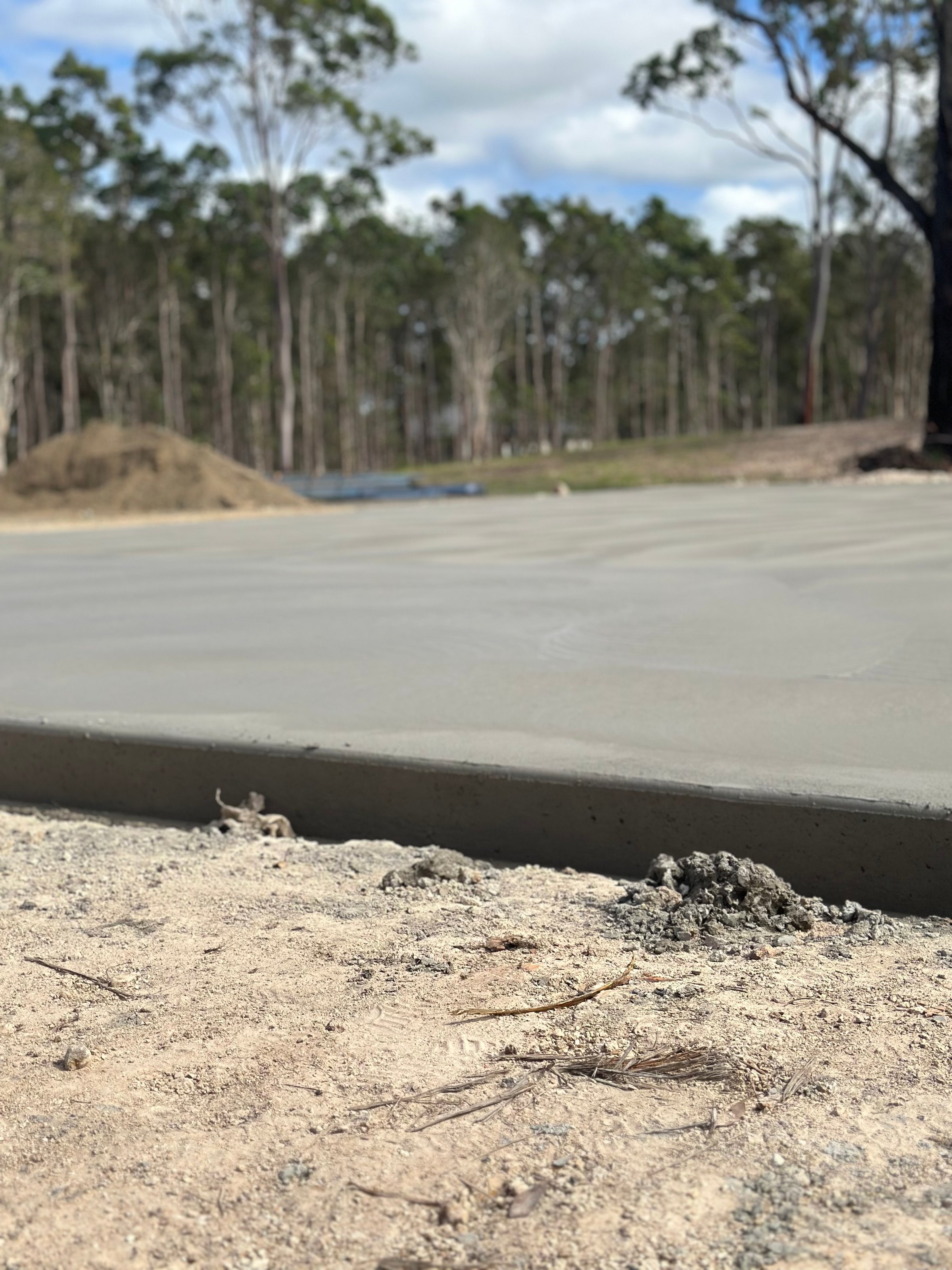 A freshly poured concrete slab surrounded by dirt and trees, with a forested background and clear sky.