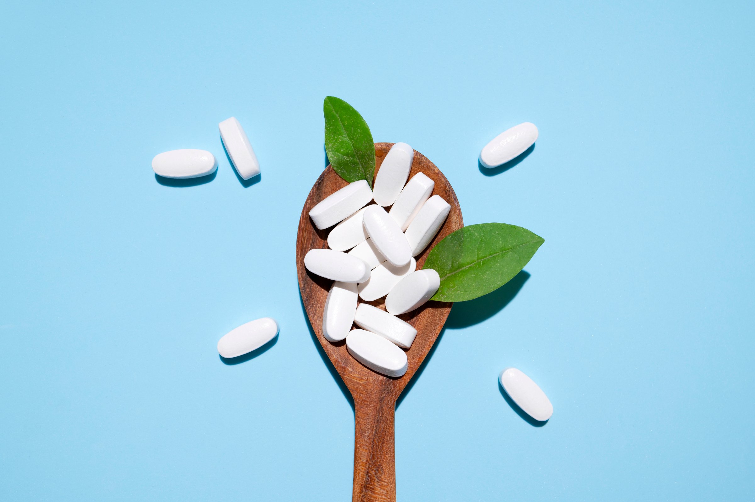 White tablets, vitamins, pills and green leaf in wooden spoon on blue background top view. Medical pharmaceutical background, health concept, organic dietary supplements, self care