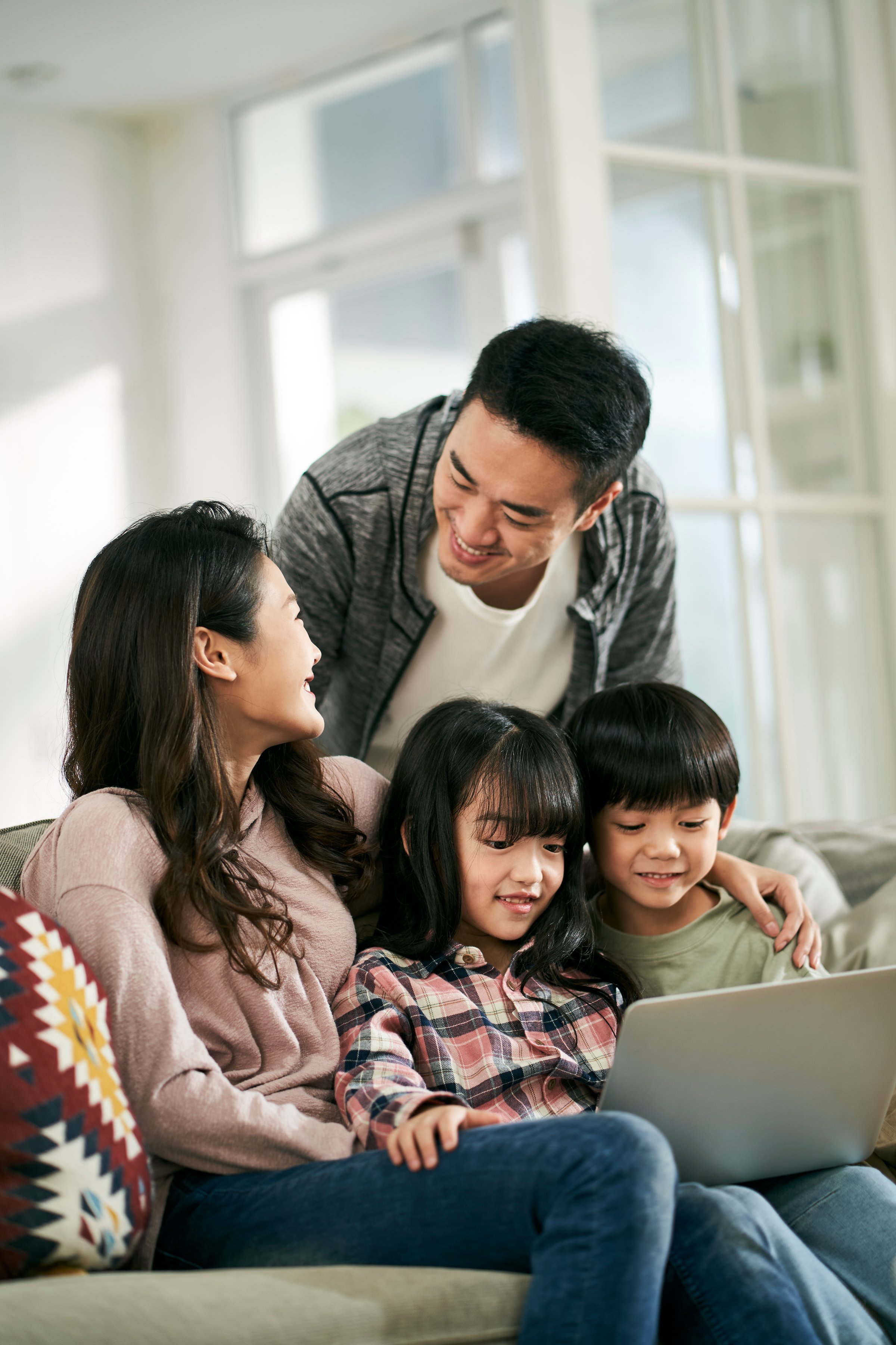 young asian couple and two children sitting on family couch at home watching movie together using laptop computer