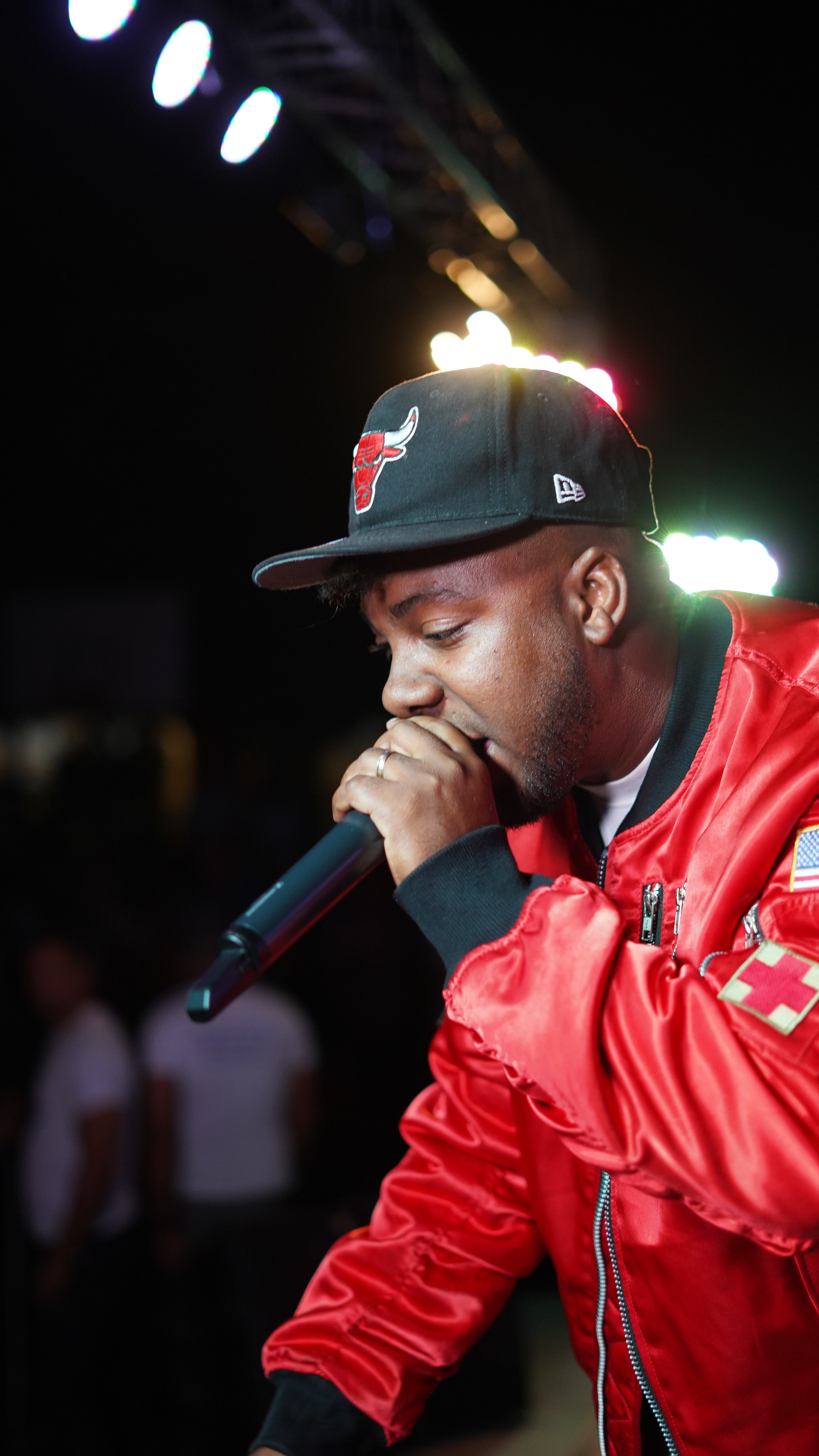 Man in a red jacket and cap holds a microphone while performing on stage under colorful lights.