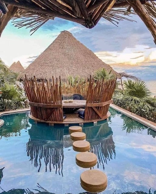 Tropical setting with a round, thatched-roof hut surrounded by a pool and stepping stones, lush greenery, and a scenic view.