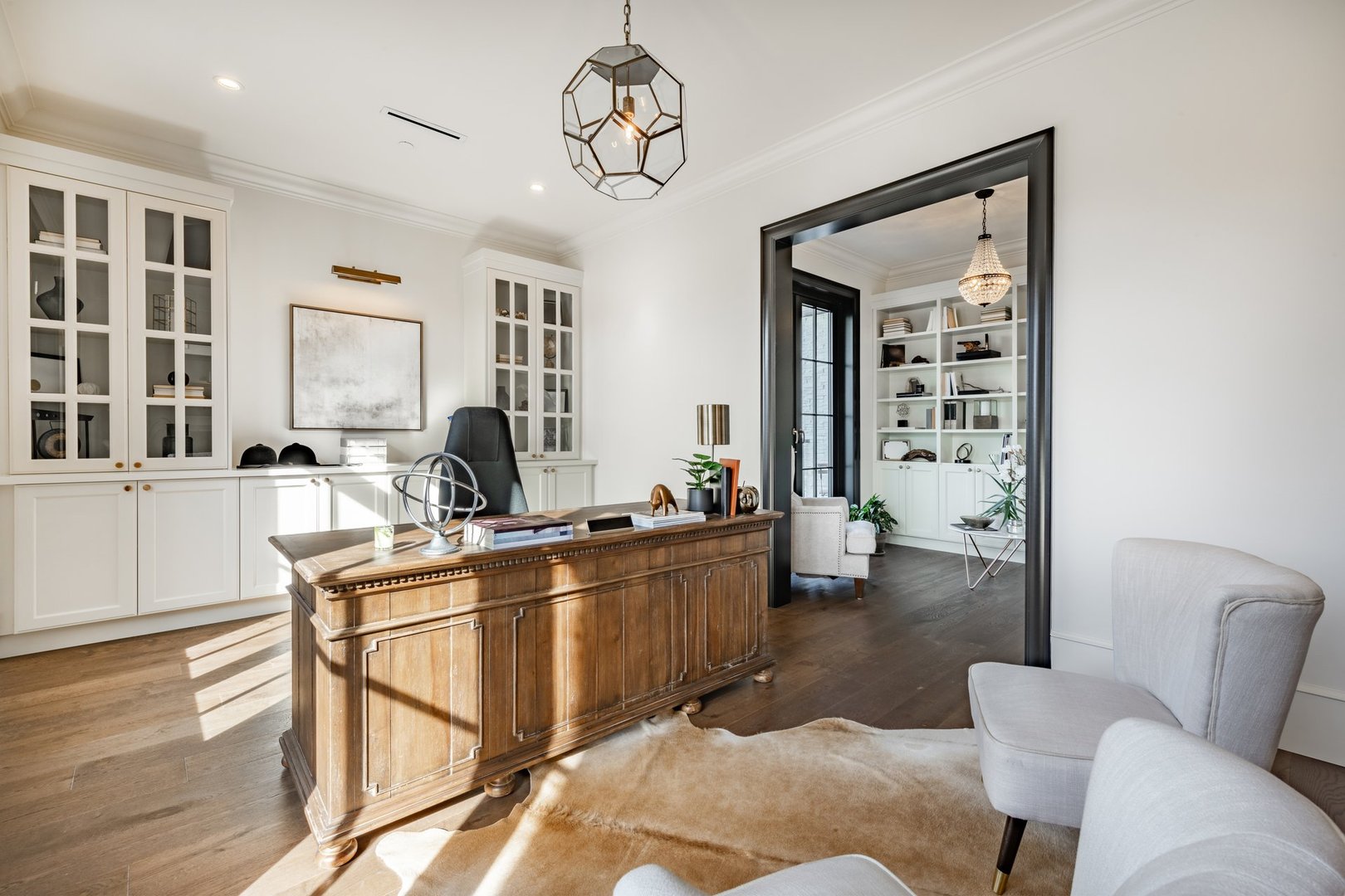 A very large home office with a wooden desk armchairs office chair built in shelving reading room attached there is a chandelier and an animal hide skin rug on the hardwood floor. A very elegant and stately workspace den study library