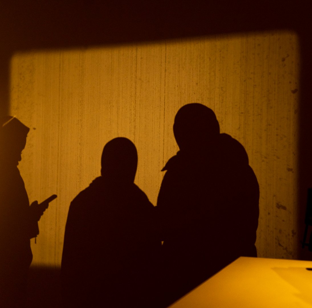 Three people are standing in a dimly lit room with a yellow light shining on the wall behind them. The image is a silhouette of the individuals, creating a sense of mystery.