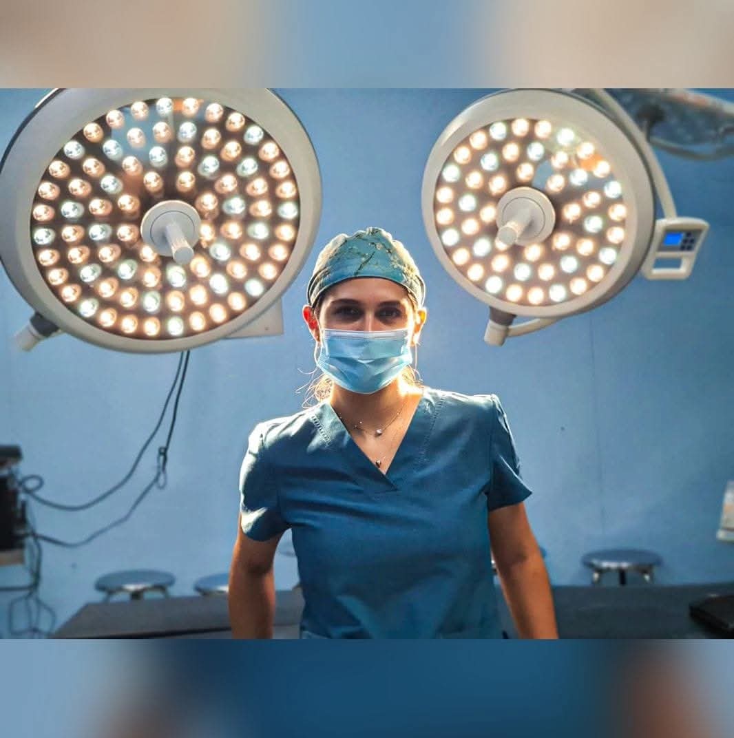 A medical professional in scrubs and a mask stands under bright surgical lights in an operating room.