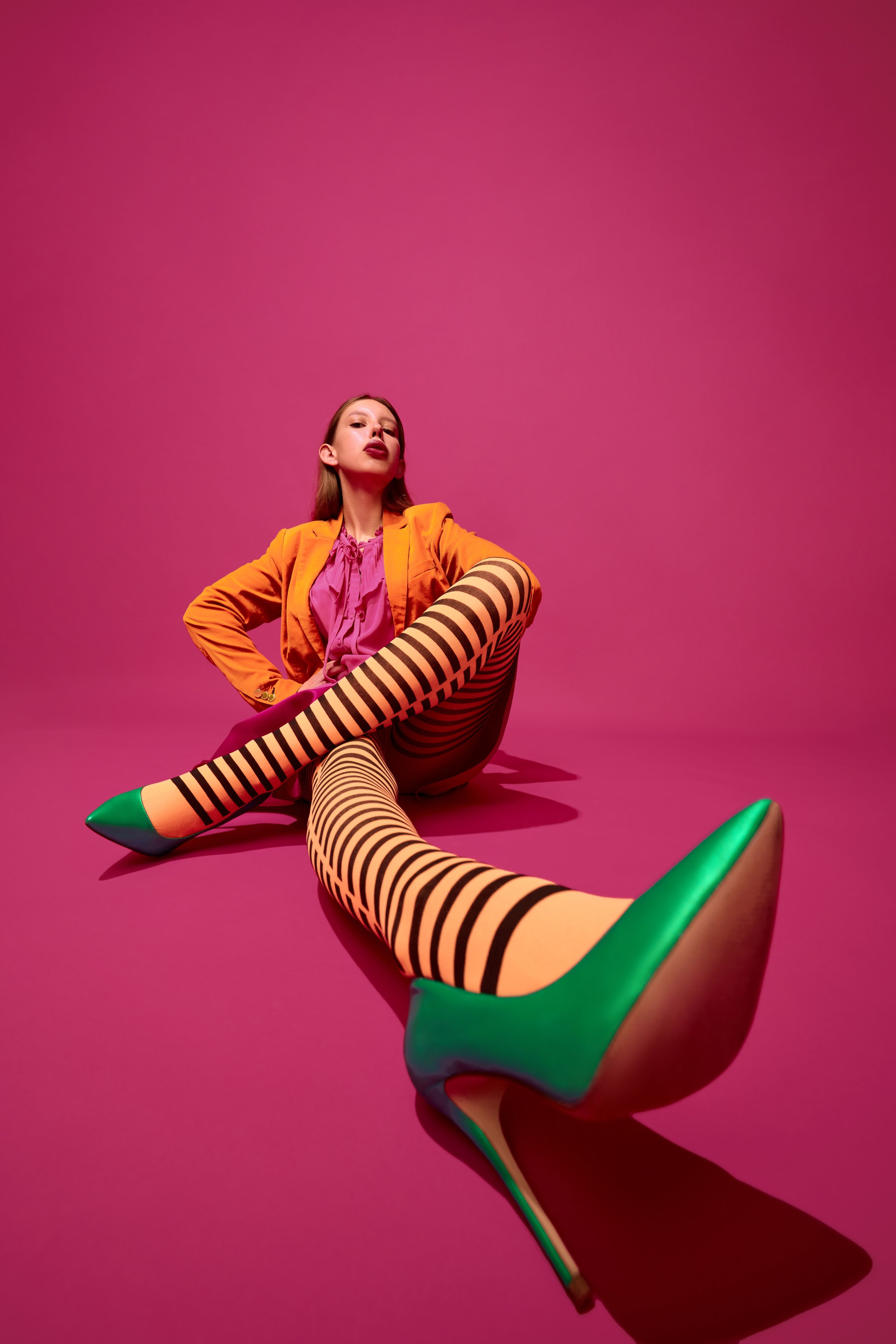 Creative full length portrait of expressive young girl, lady in unusual, variegated orange striped tights, long skirt, short jacket and green heels. Concept of surrealism, art, high fashion, beauty.