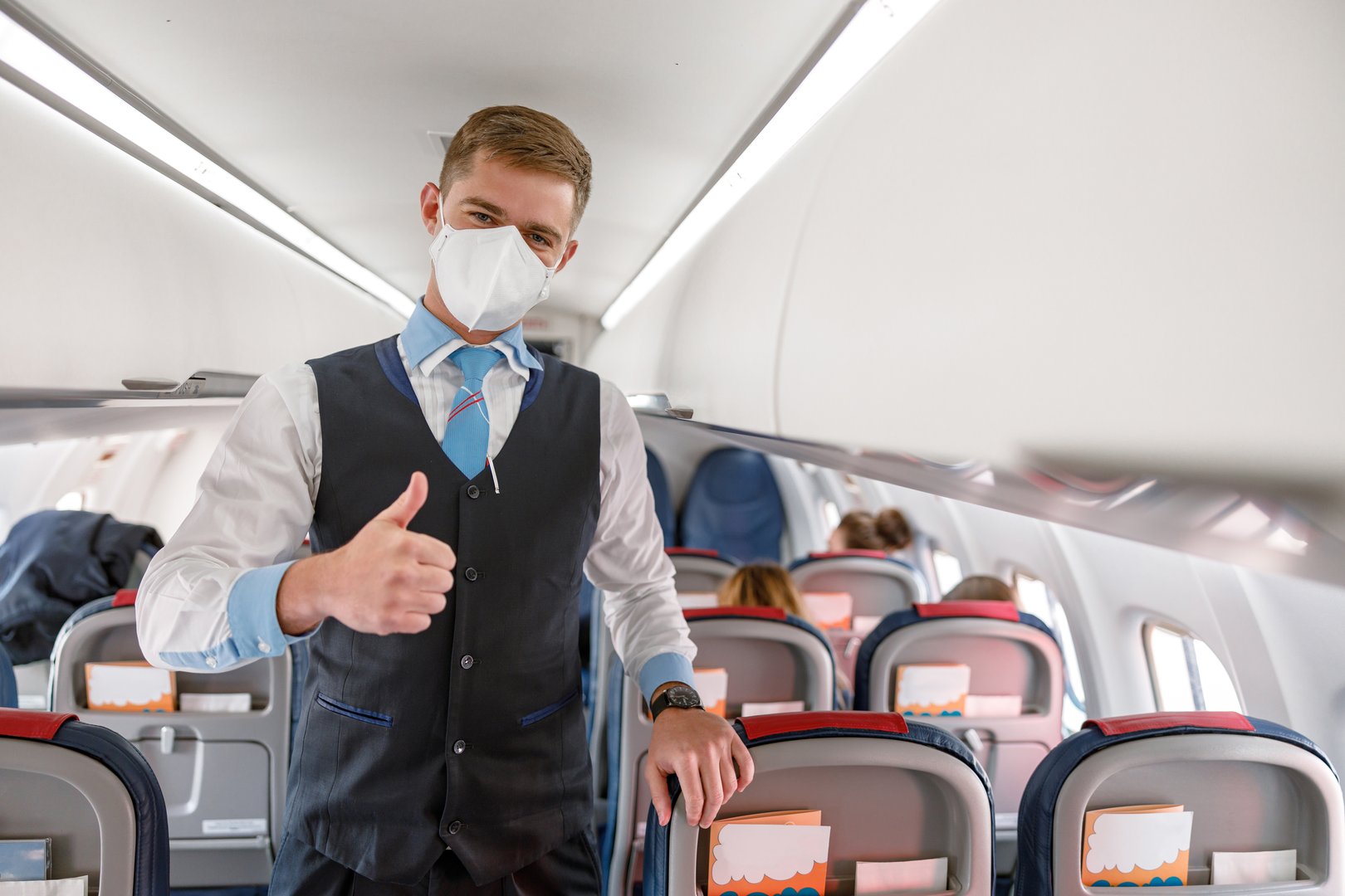 Man flight attendant in protective face mask standing near passenger seat and shoving approval gesture