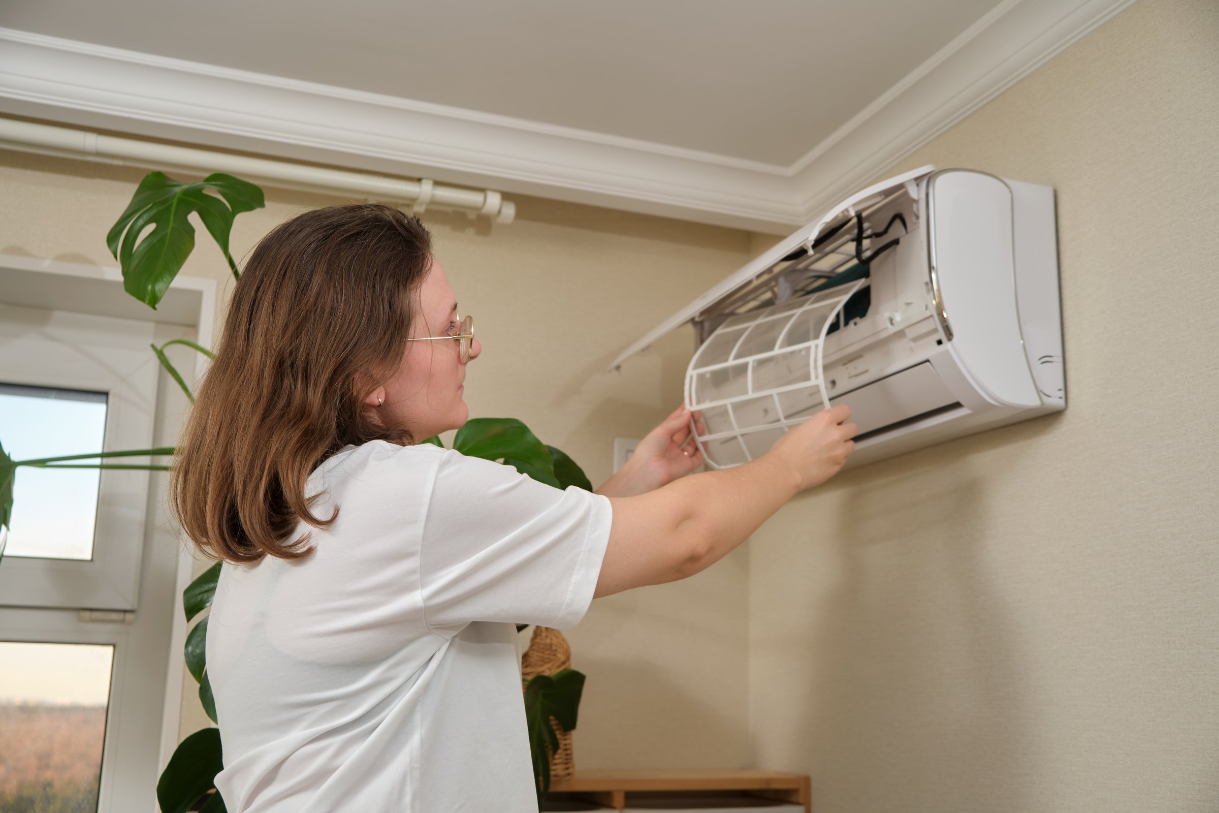 Cleaning air conditioner filter. Woman removing dust filter from wall-mounted split air conditioning unit for maintenance. Home appliance care and HVAC system maintenance.