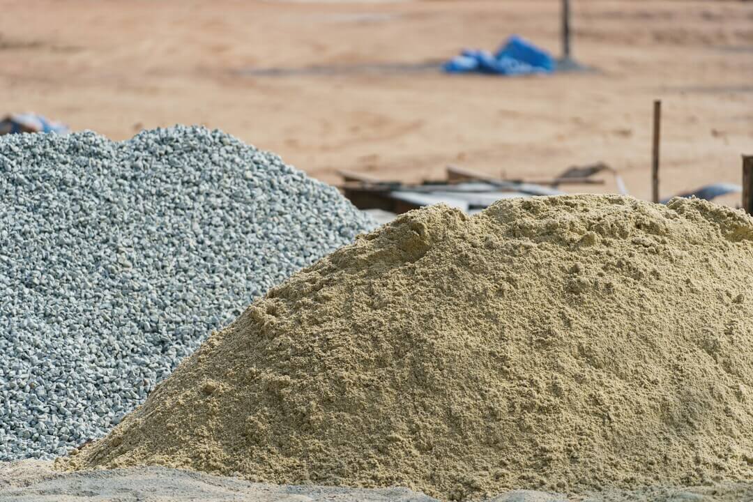Piles of gravel and sand at a construction site with a blurred background of sandy ground and scattered materials.