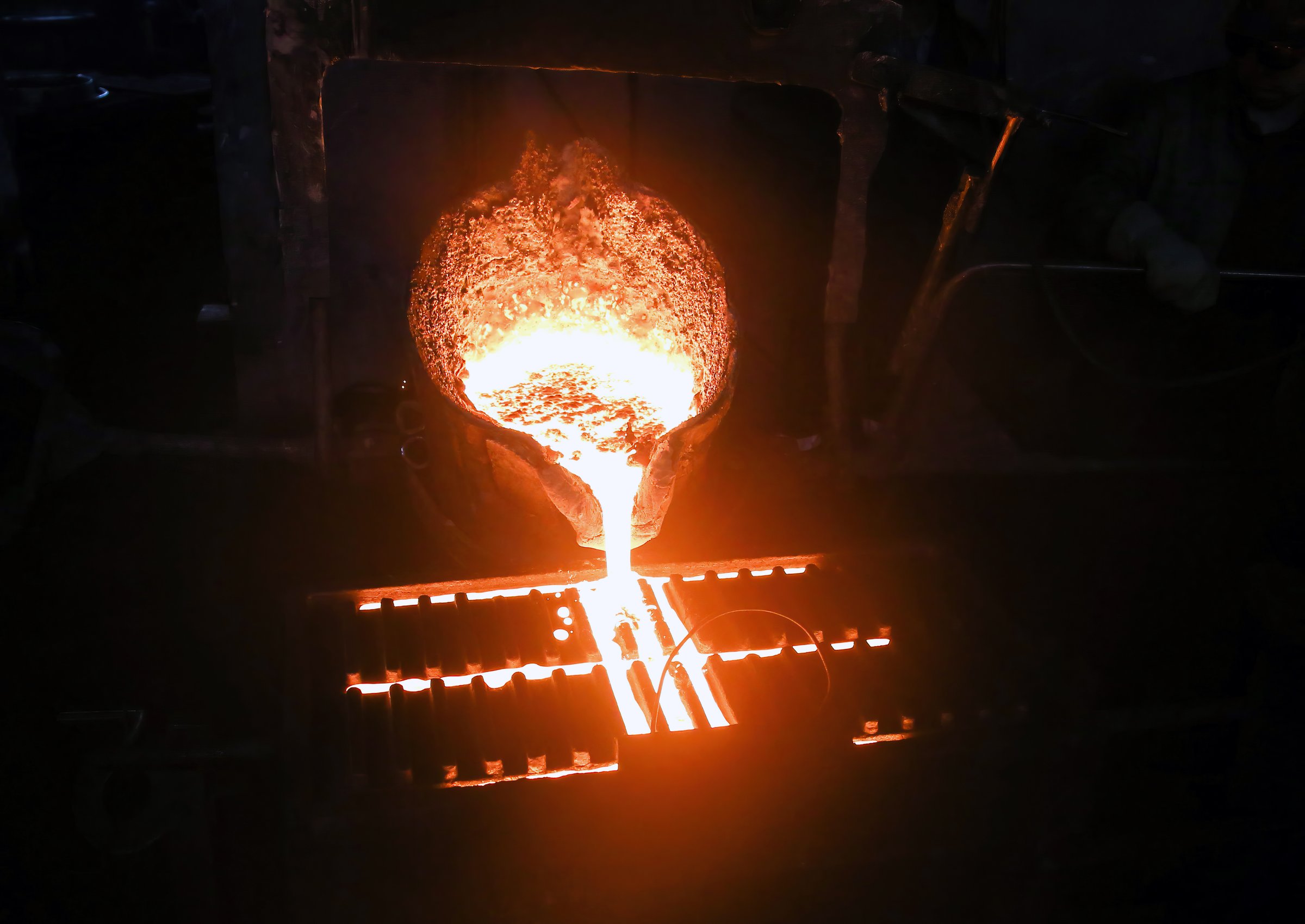 Molten metal poured from ladle into mold to make casting. Metal glows brightly, flows into mold, sparks fly as metal hits mold, on dark background, showcasing metal casting process.