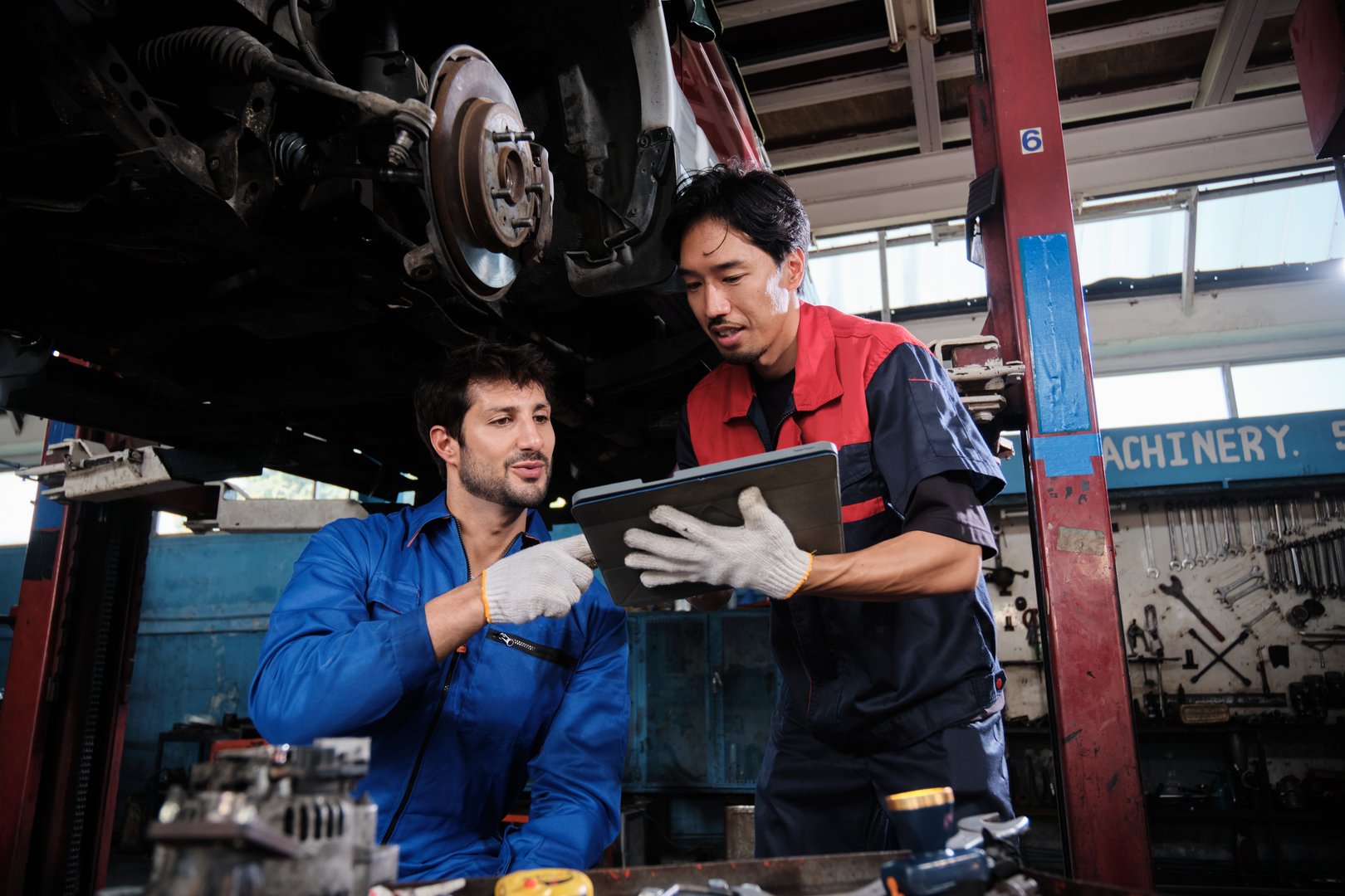 Two male mechanics cooperate, Latin and Asian workers fixing automotive wheels and maintaining car suspensions in garage station, and technician engineering service jobs in vehicle transport industry.