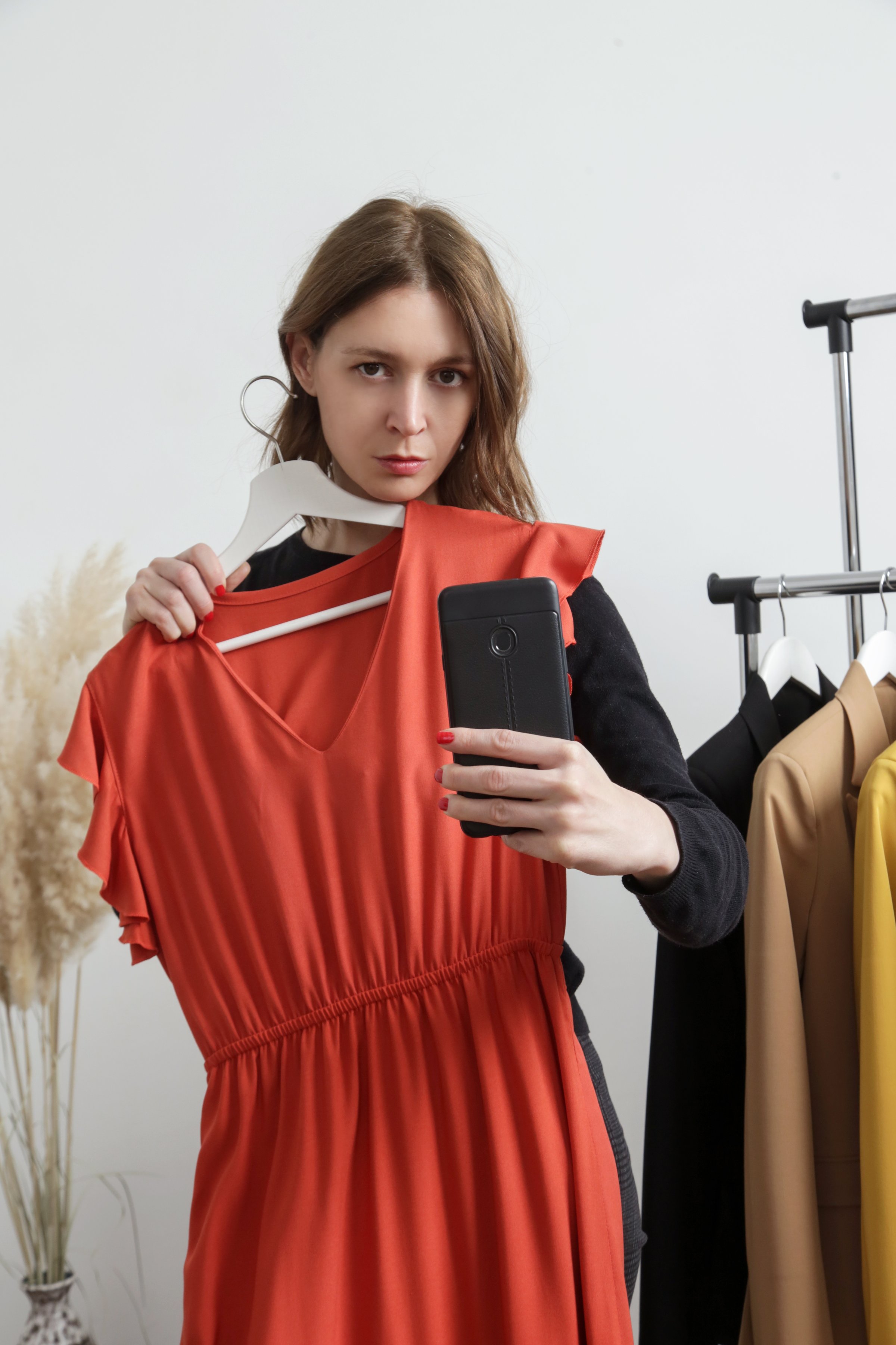 Beautiful young woman near rack with clothes holding dress and taking mirror selfie.