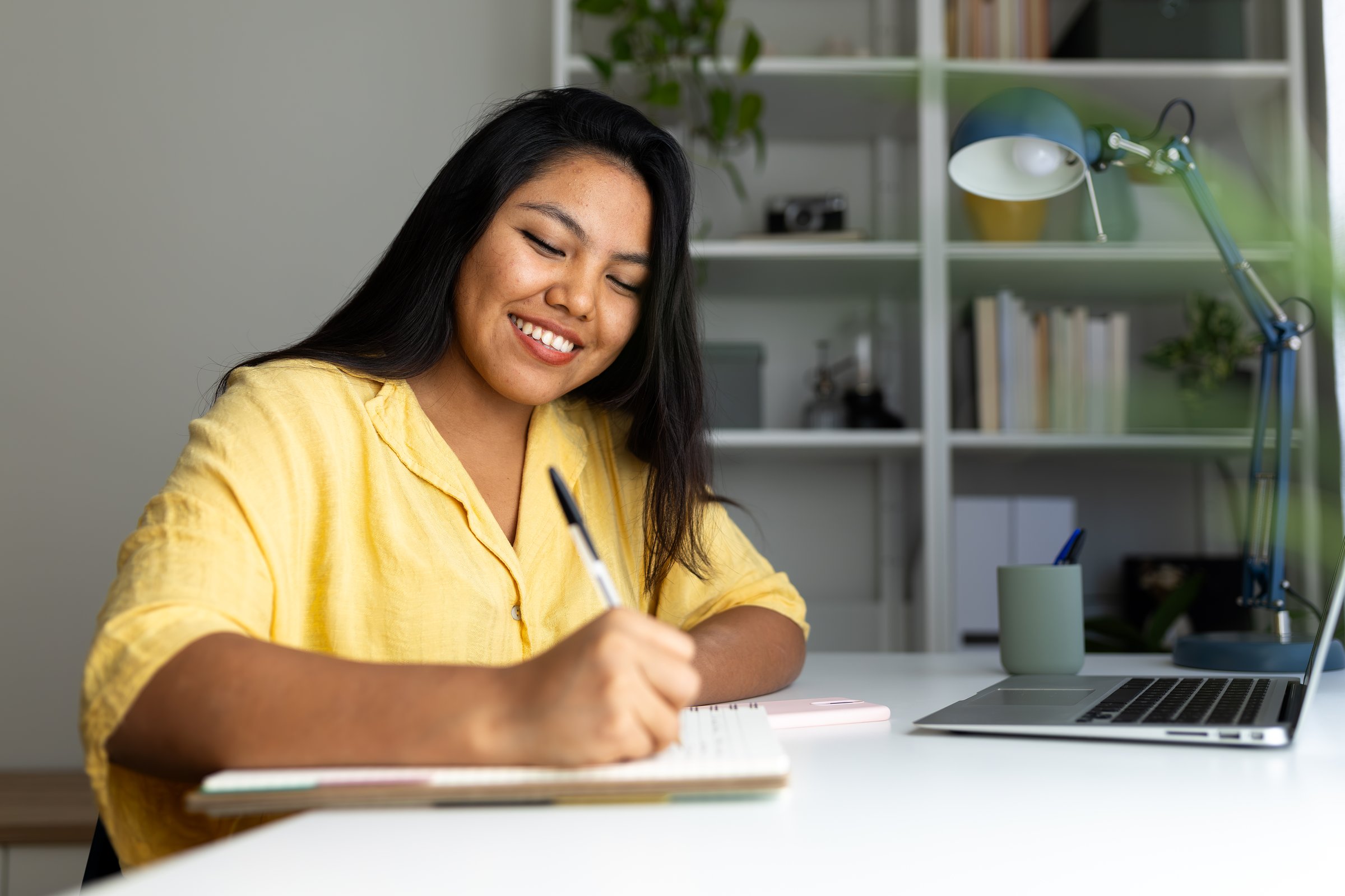 Young latin woman writing on notebook and smiling while working from her home office