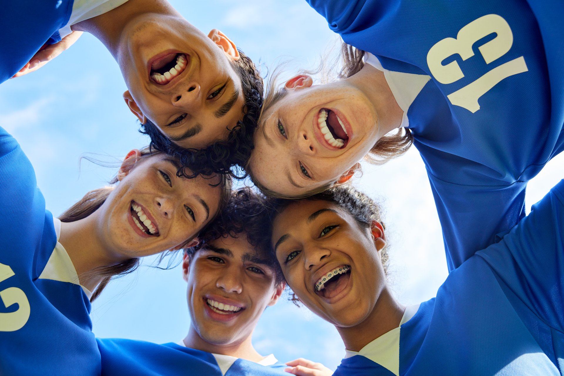Portrait of young sports team in a close huddle smiling. Group of guys and girls forming a circle with happy faces before match. Cheerful multiethnic teammates looking at camera in unity while they hug each other and touch their heads.