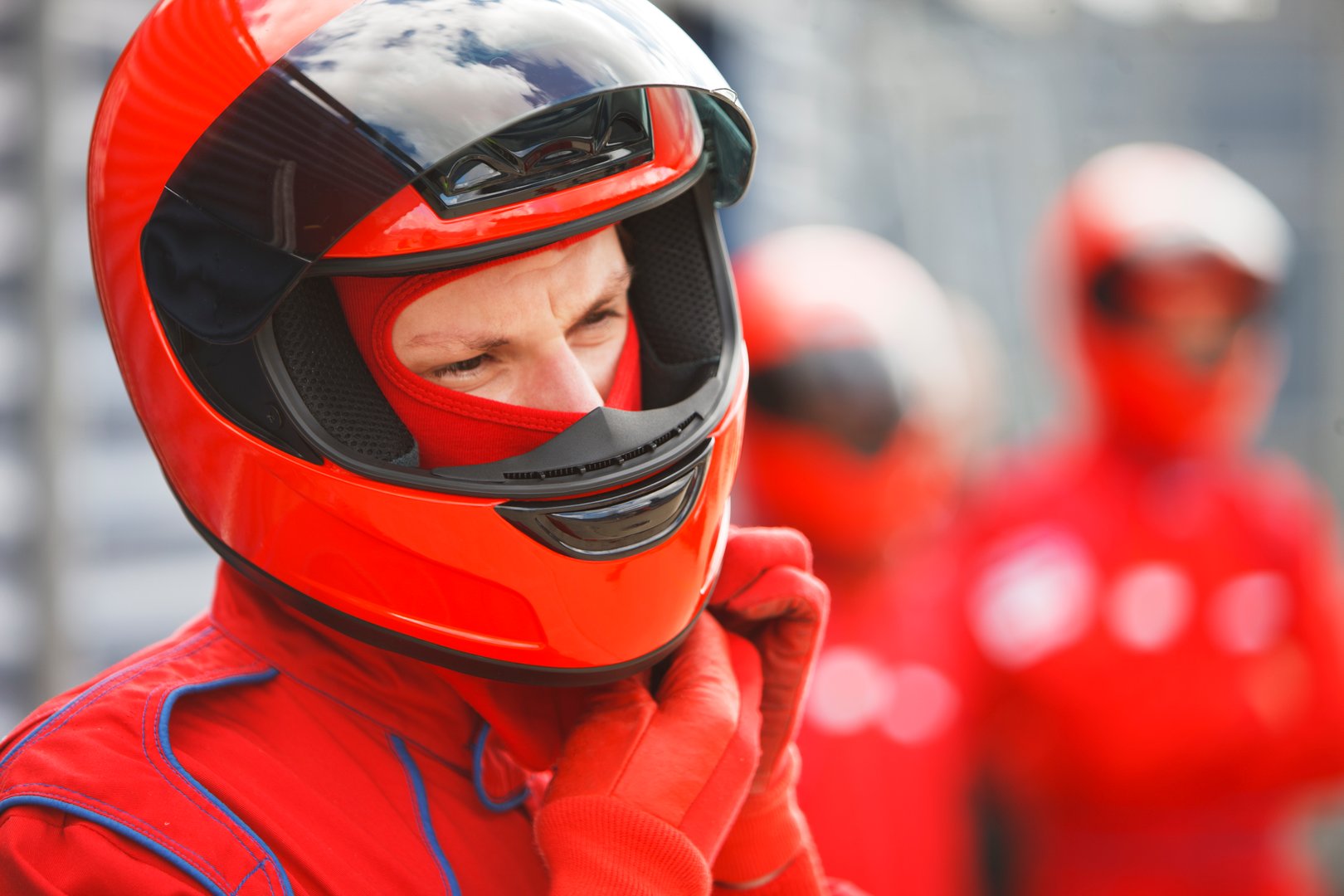Racecar driver in a red racing suit and helmet, adjusting chin strap, with blurred team members in the background.