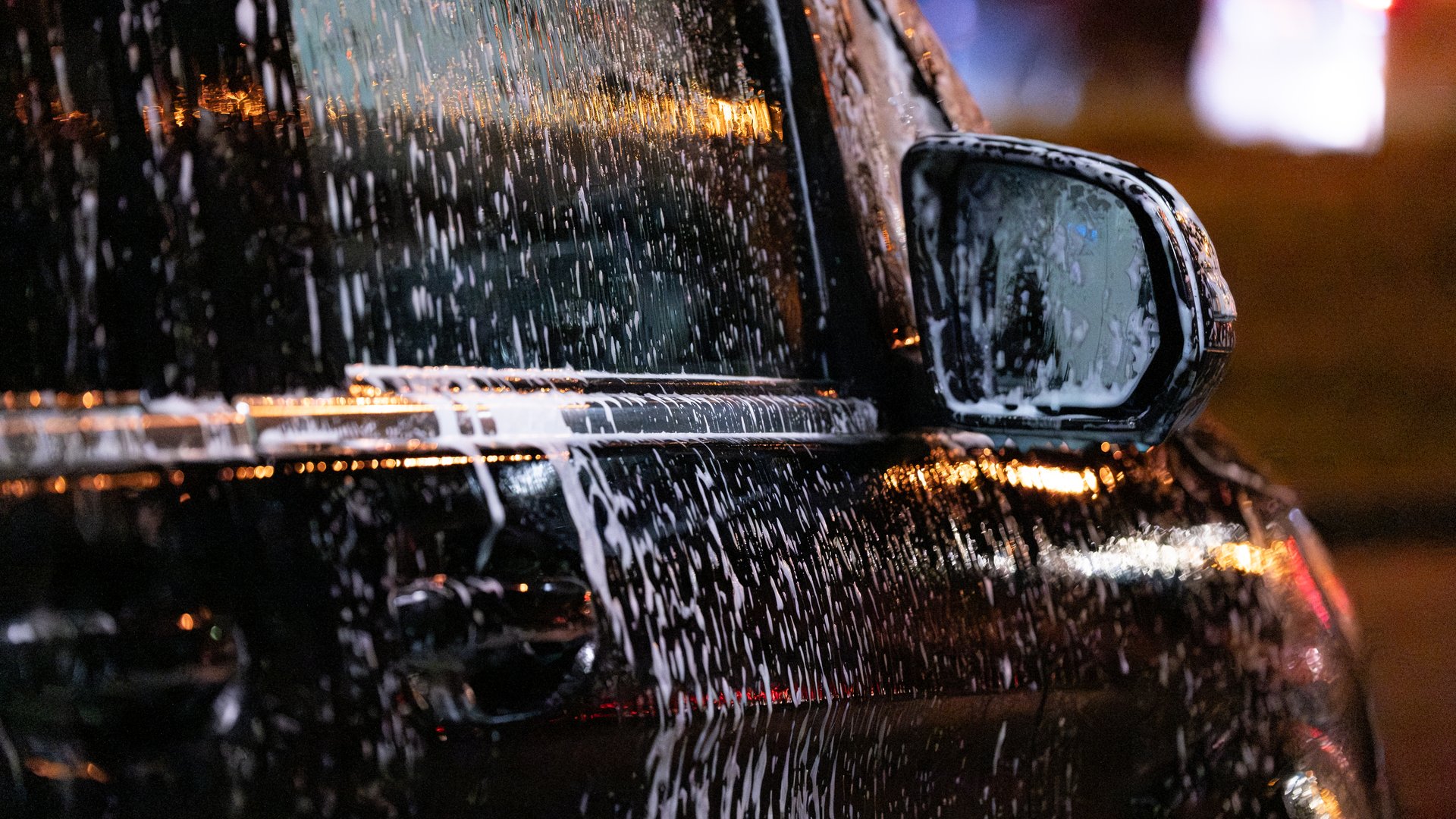 Close-up of a black car covered in soap and water during washing at night. Reflections of city lights highlight the glossy surface and foam texture. The image represents auto care, detailing, and modern urban lifestyle. Ideal for illustrating car wash services or vehicle maintenance.