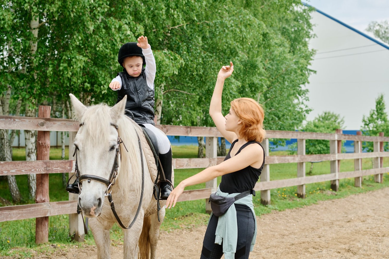 Young instructor guiding child in mastering horseback riding skills outdoors, with verdant trees and wooden fence creating serene backdrop for learning experience