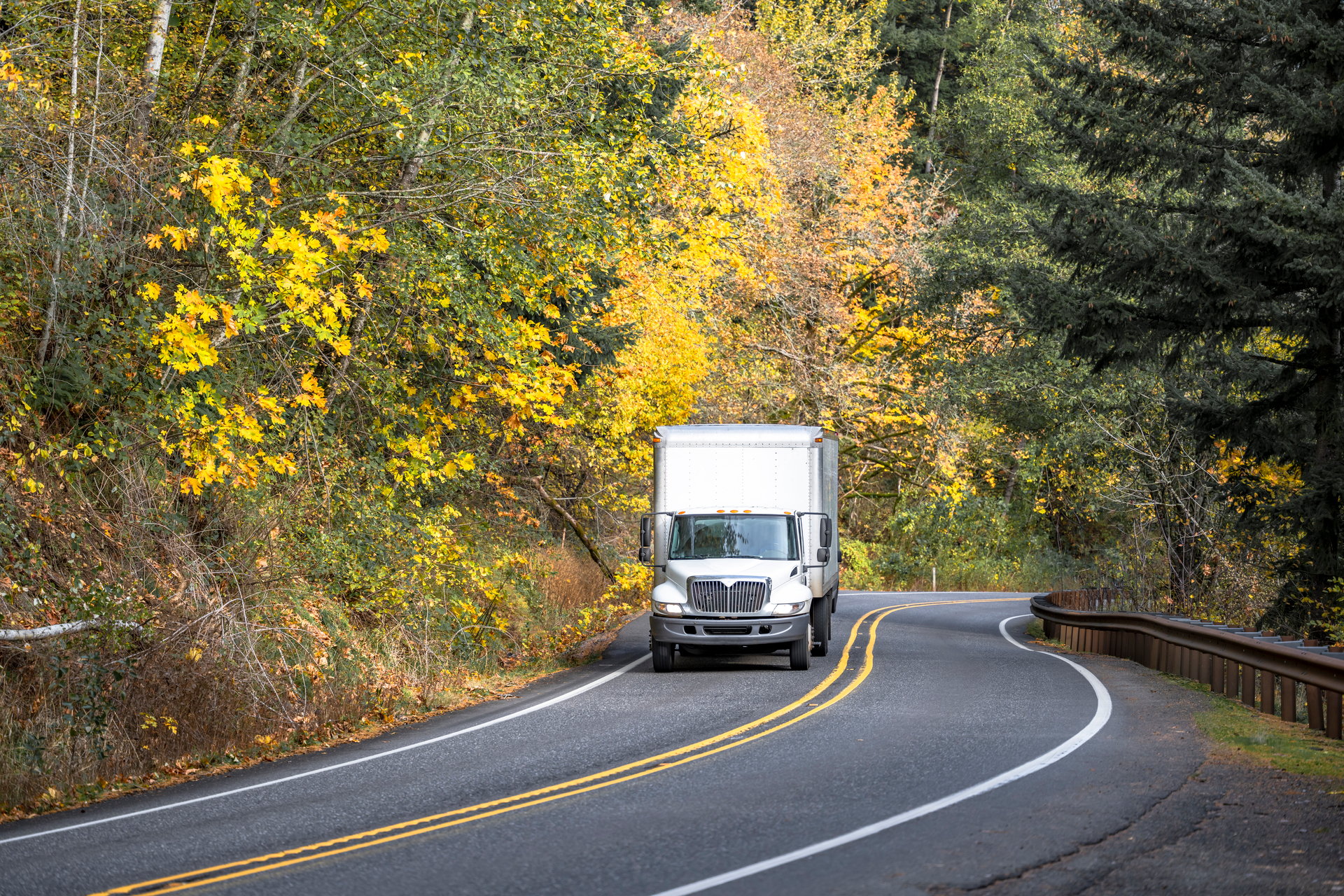 Industrial medium power day cab white rig semi truck transporting commercial cargo in long box trailer driving on the winding narrow road through the autumn forest in Columbia Gorge area