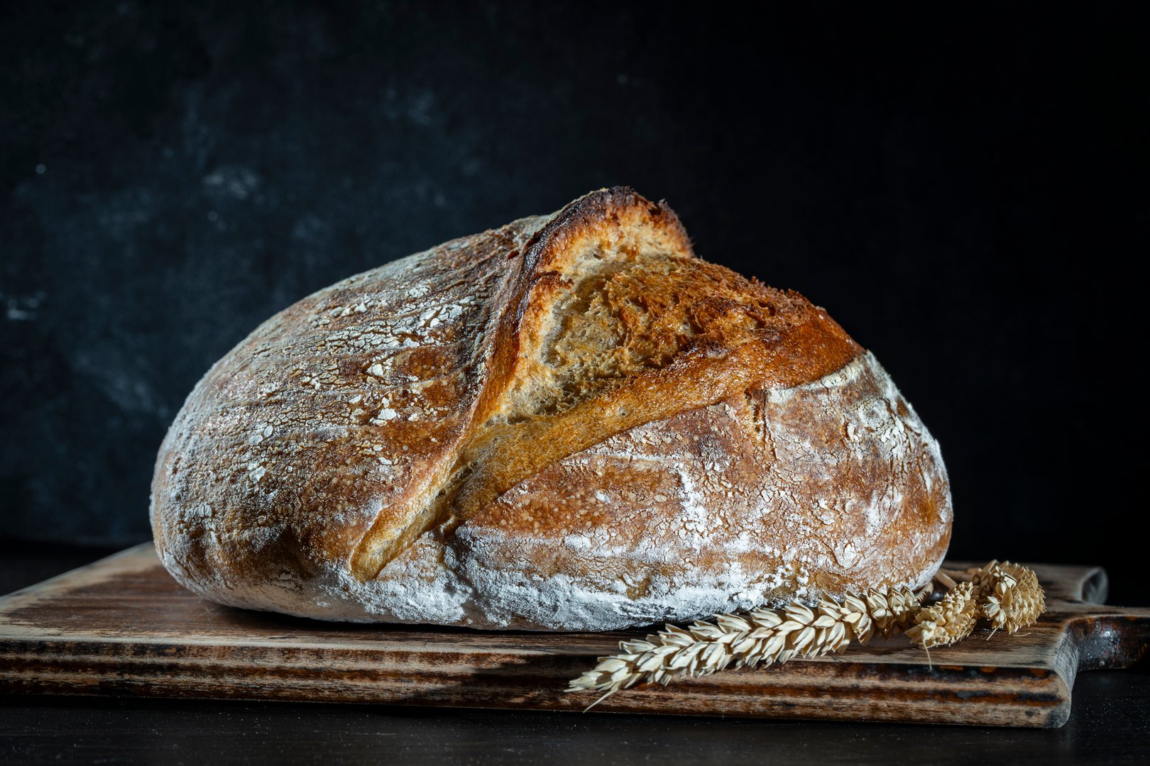 Round loaf of freshly baked sourdough bread with ears of wheat on wooden cutting board, close up. Rustic sourdough bread. Cooking healthy sourdough bread at home