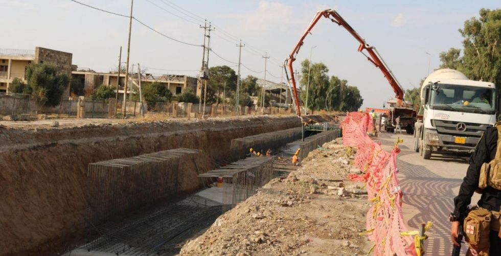Construction site with concrete mixer and workers near a trench, rebar framework visible, safety barriers in place.