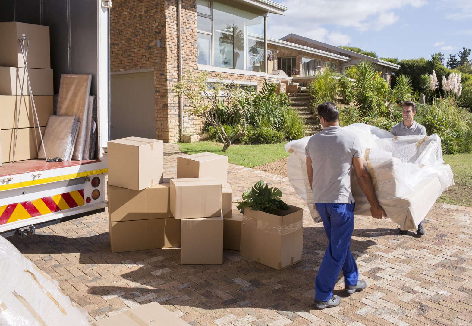 Two movers carrying furniture wrapped in plastic, with cardboard boxes around, near a house and a moving truck.