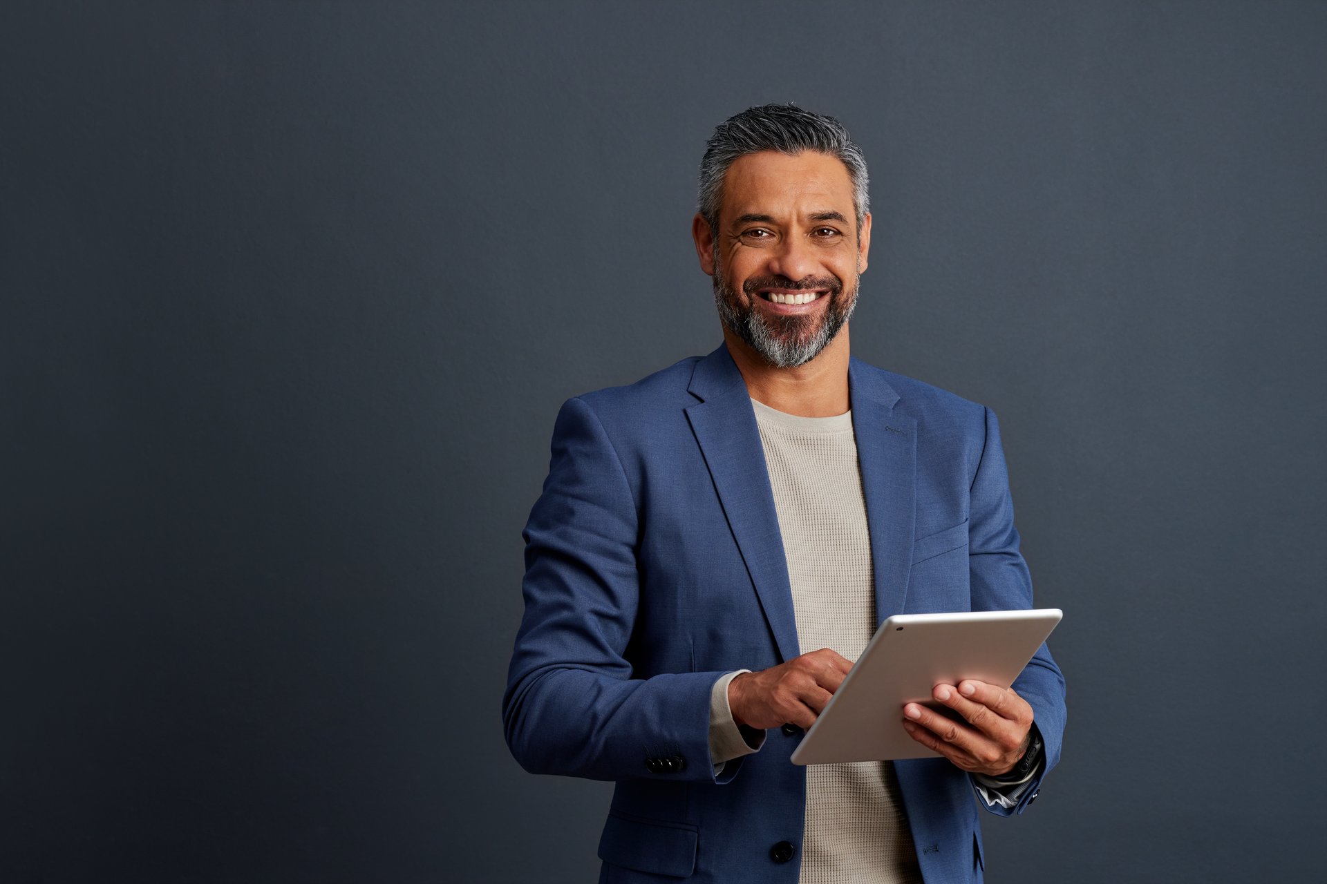 Smiling middle aged Indian man holding a digital tablet, dressed in a blue blazer. Successful middle aged business man using a tablet, standing against a gray background with copy space.