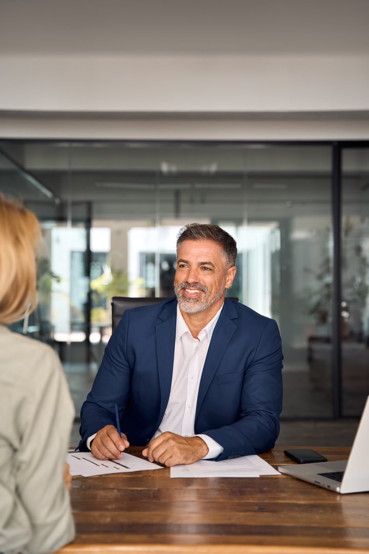 Lawyer or real estate agent make consultation for client. Mature Latin business man and European business woman discussing project on laptop sitting at table in office. People work together, vertical.