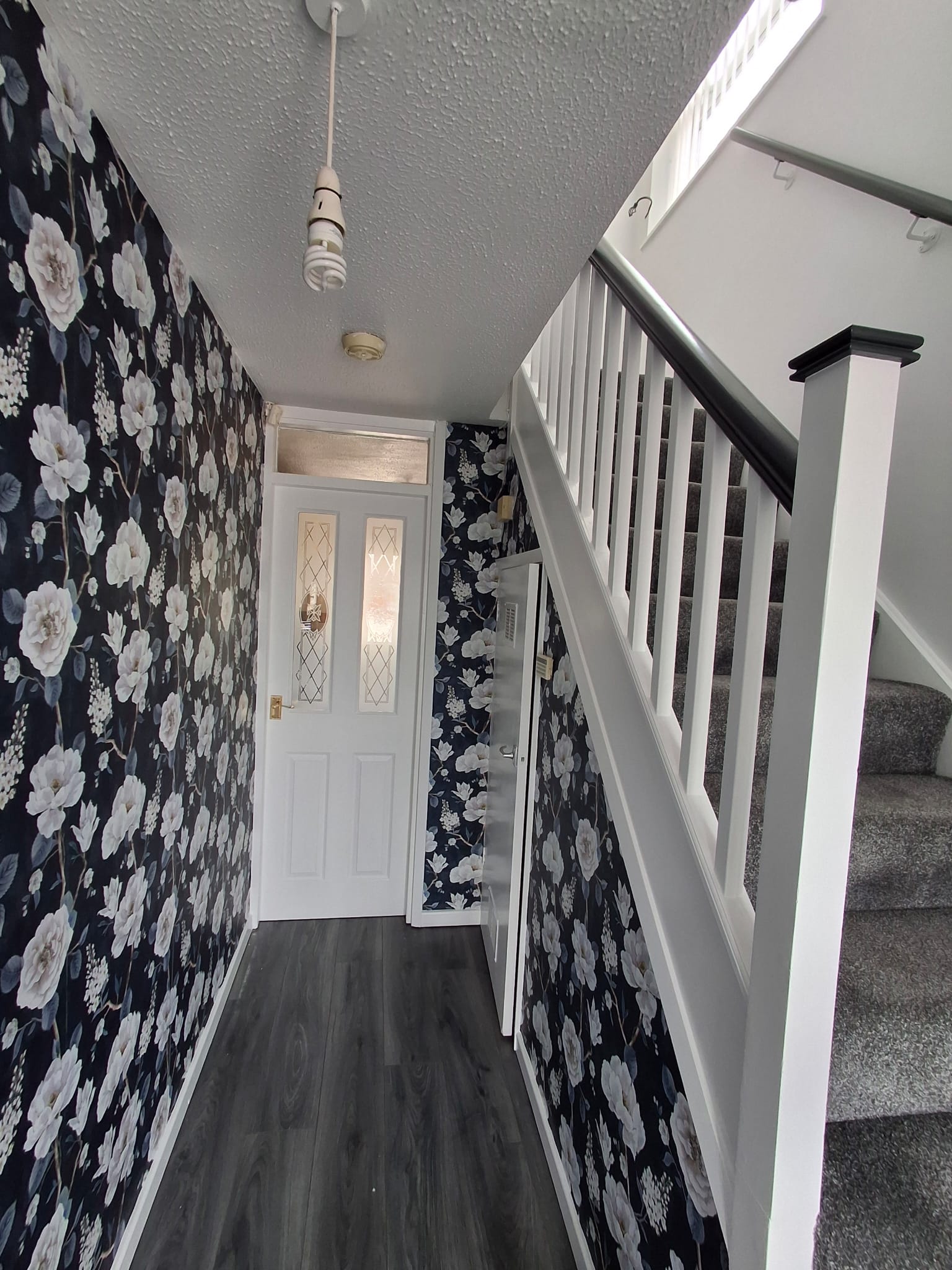 A hallway with dark wood flooring, floral wallpaper, a white door with glass panels, and carpeted stairs with a white railing.