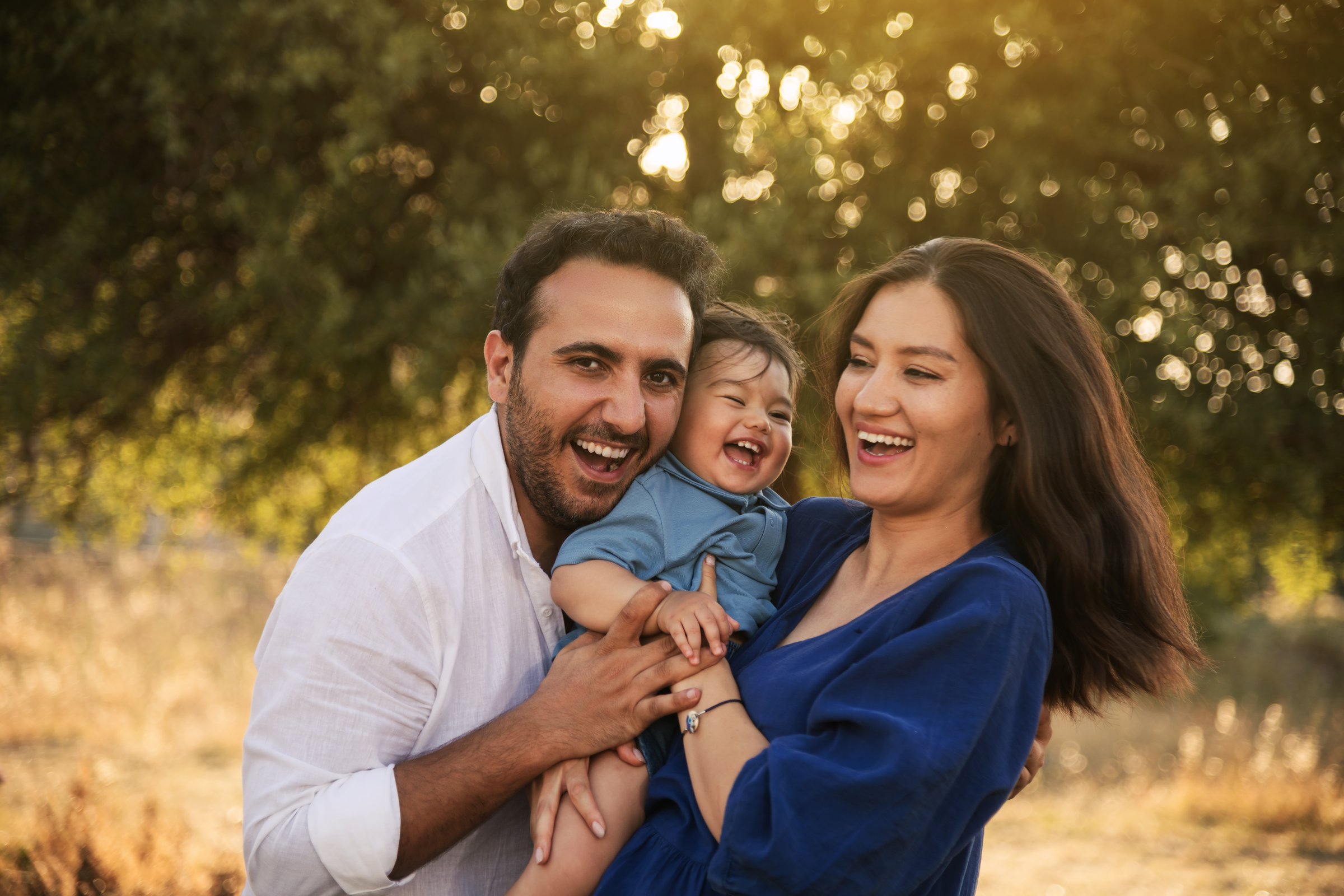 A joyful family moment with a father, mother in a blue dress, and their one-year-old son in blue clothes, hugging and smiling in a grassy natural setting with a tree in the background