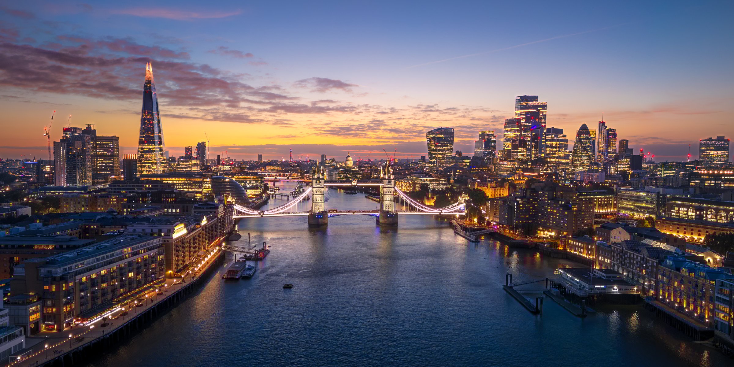 A stunning aerial panoramic view of London at twilight, featuring the Tower Bridge illuminated over the River Thames and the modern City skyline glowing in the background. Captures the vibrant energy, architecture, and beauty of London as day transitions to night.