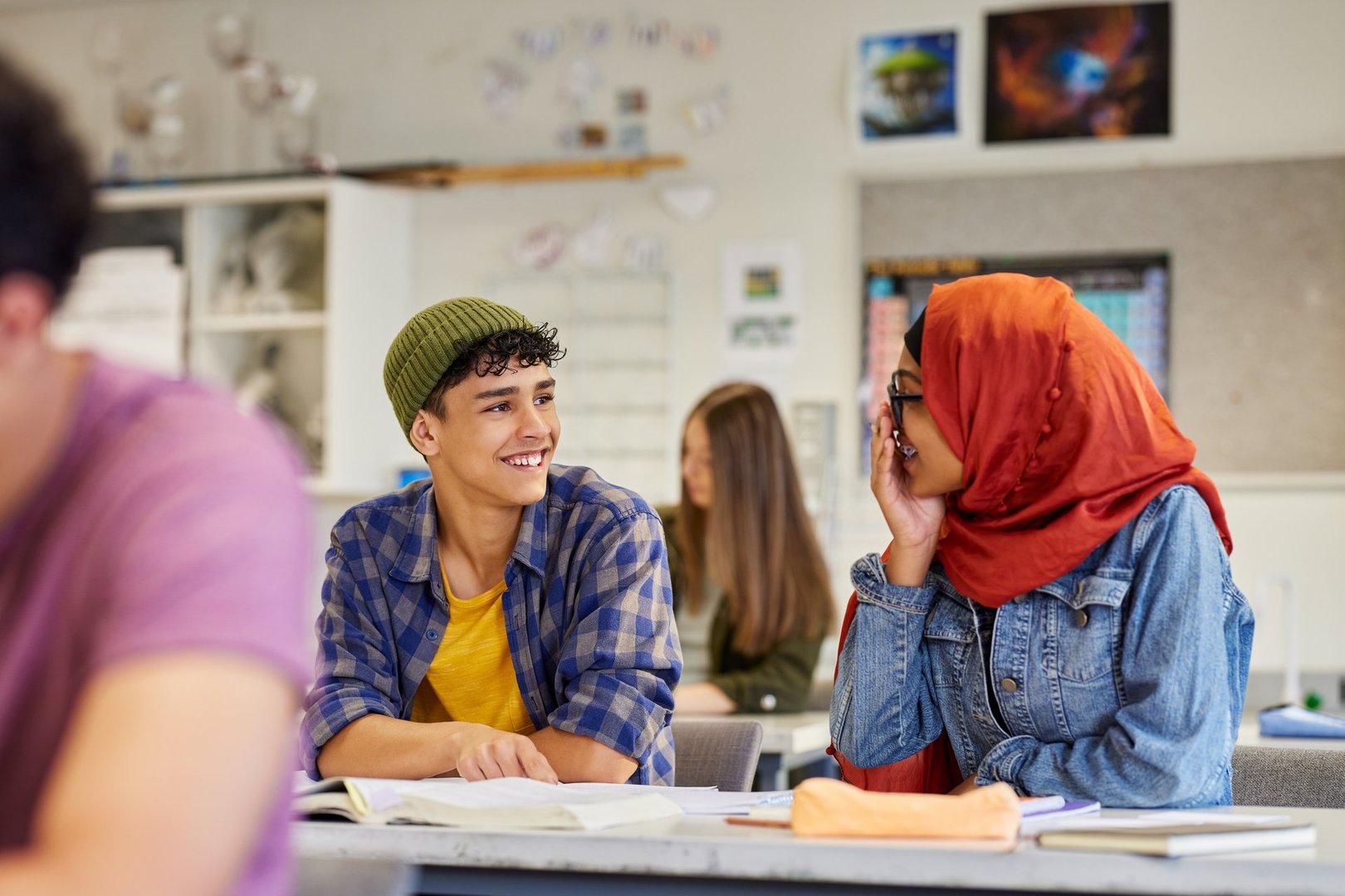 Two students smiling and talking happily during lesson at secondary school. Latin guy and islamic girl studying together in class. Boy and girl with hijab communicating and joking with each other during test in classroom at high school.