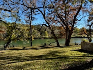 A peaceful riverside scene with trees, a grassy lawn, and calm water under a clear blue sky.