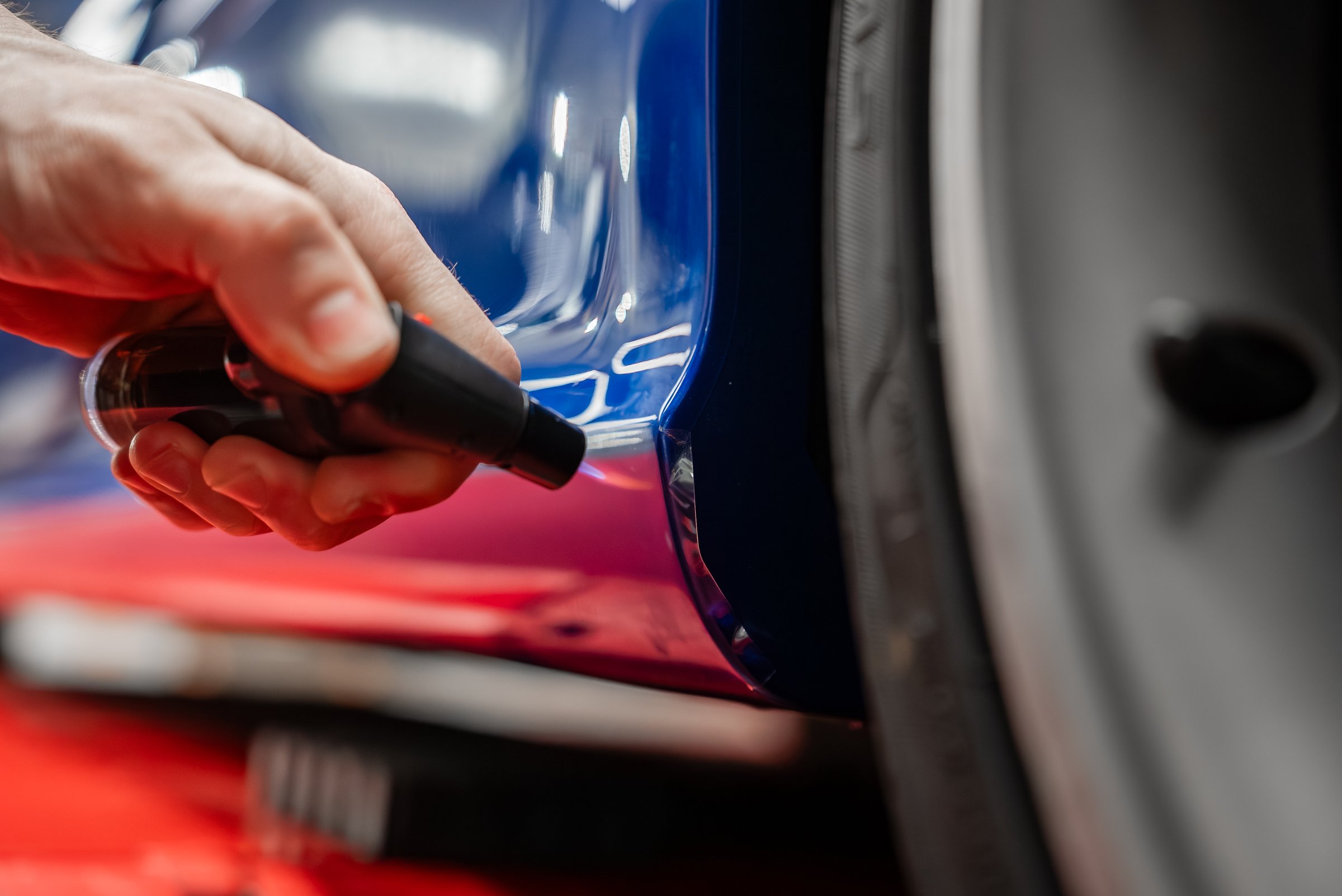 A technician applies touch up paint to a fine scratch on a glossy blue car panel beside a tire in a luxury dealership service bay, under controlled lighting and red floor.