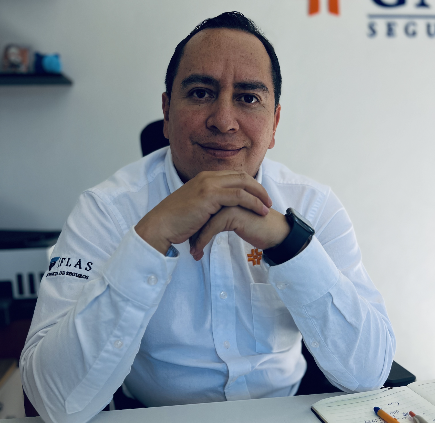 Man in a white shirt with company logos sits at a desk, hands clasped, looking at the camera.