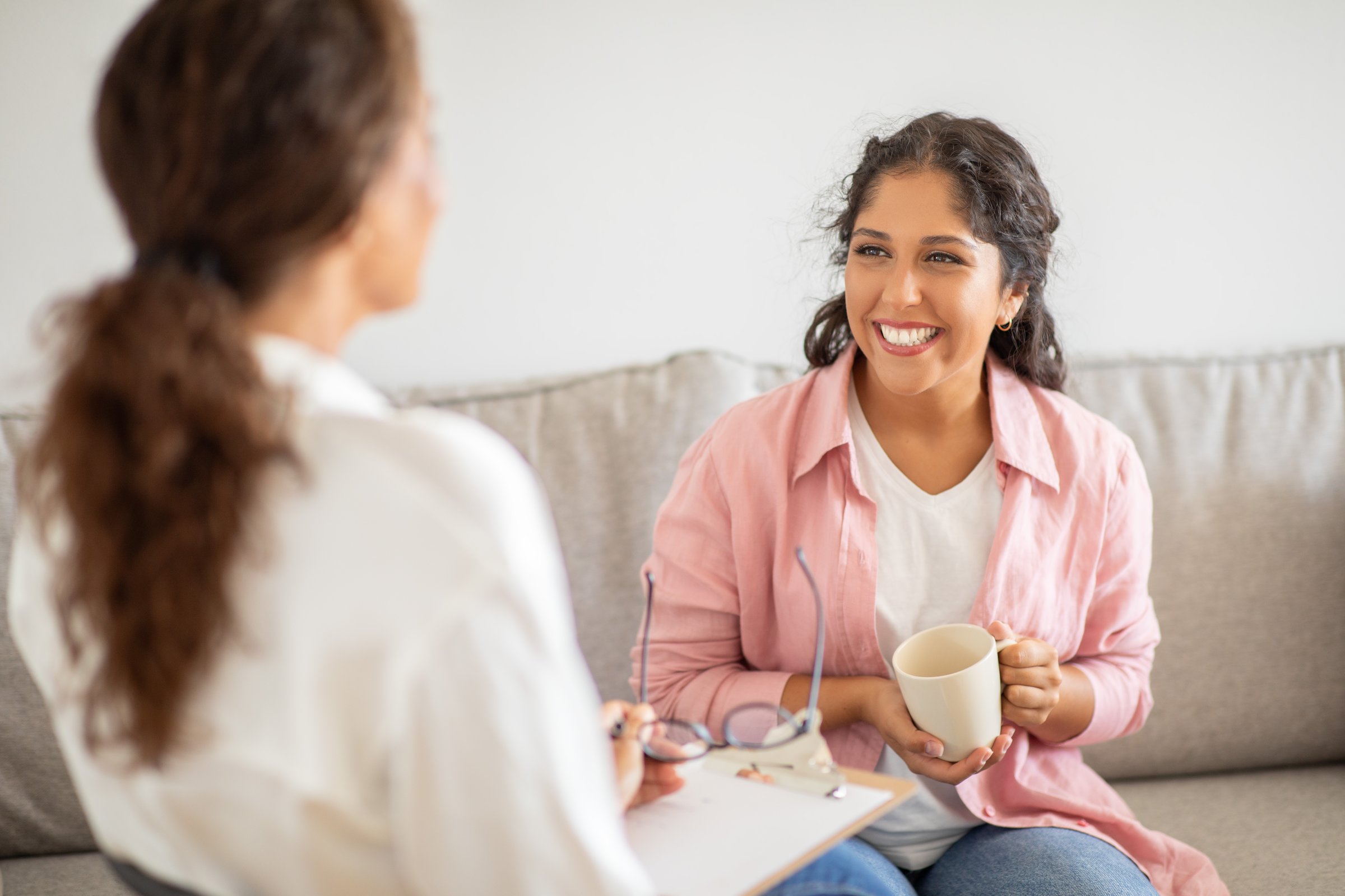 A woman wearing a pink shirt sits on a couch and smiles while holding a cup of coffee. She is talking to another woman sitting next to her, who is wearing a white shirt.