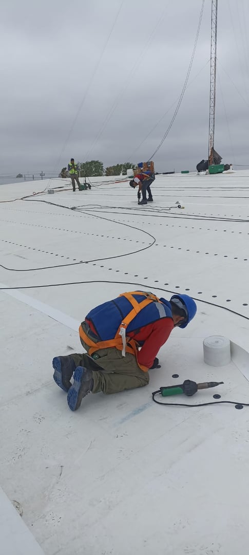Workers on a large rooftop install roofing material, wearing safety gear and using tools. Overcast sky in the background.