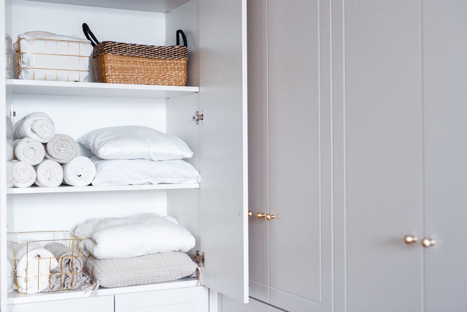 White laundry, pillows, towels organized and folded in baskets on shelves in white opened wardrobe. Clothes organization