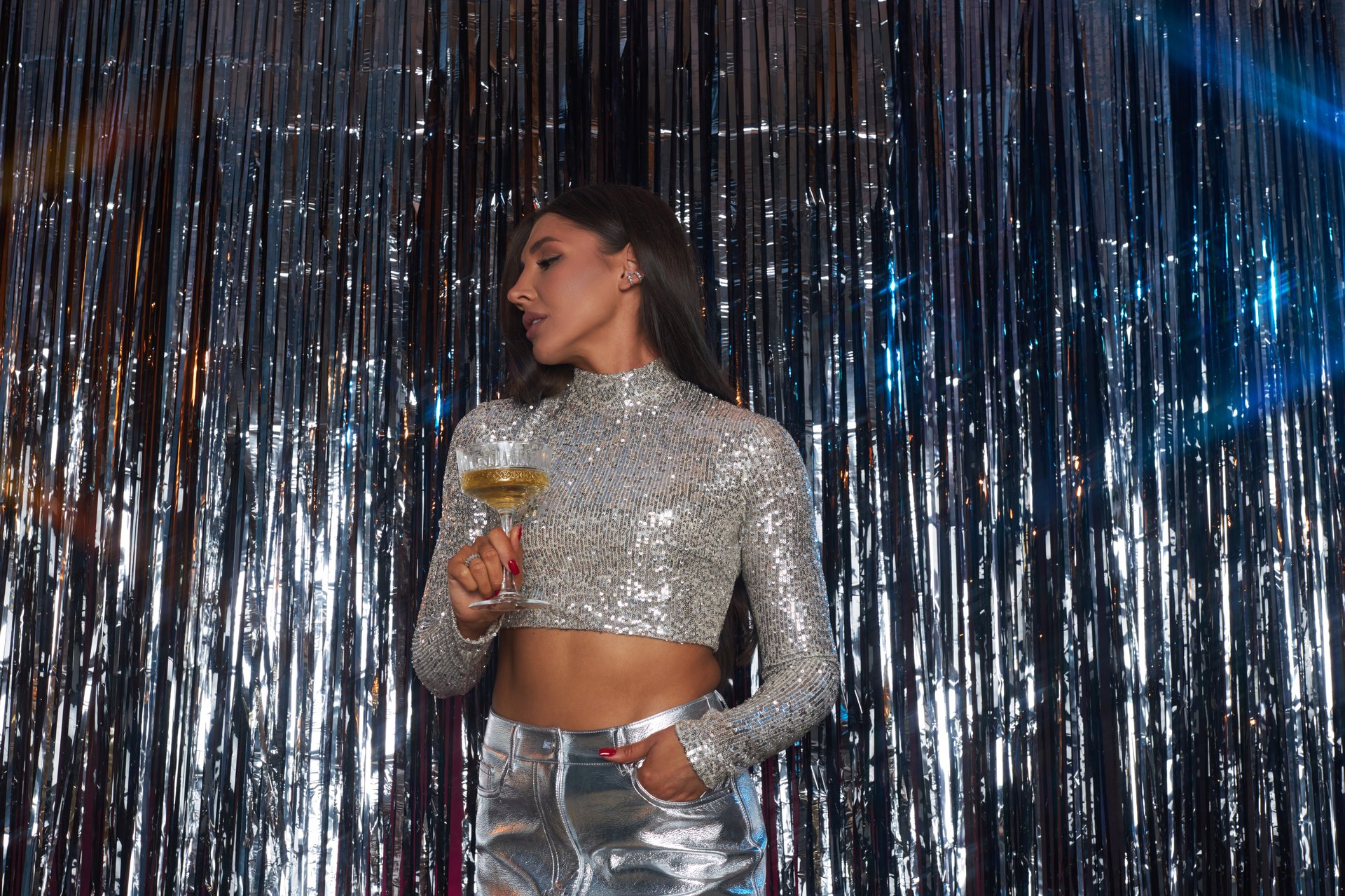 Woman standing against sparkly backdrop holding a drink in a festive environment while wearing a shimmering top. The scene suggests a lively social gathering