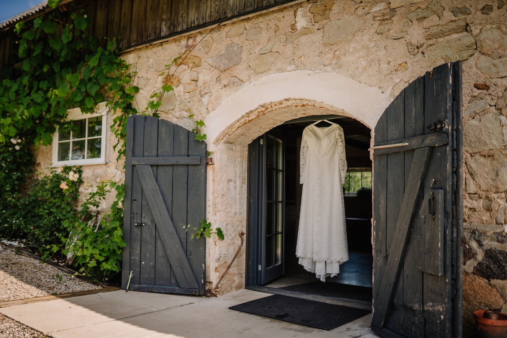 A white lace wedding dress hangs on a wooden hanger in the arched entrance of a rustic stone barn with dark blue doors and creeping green vines.