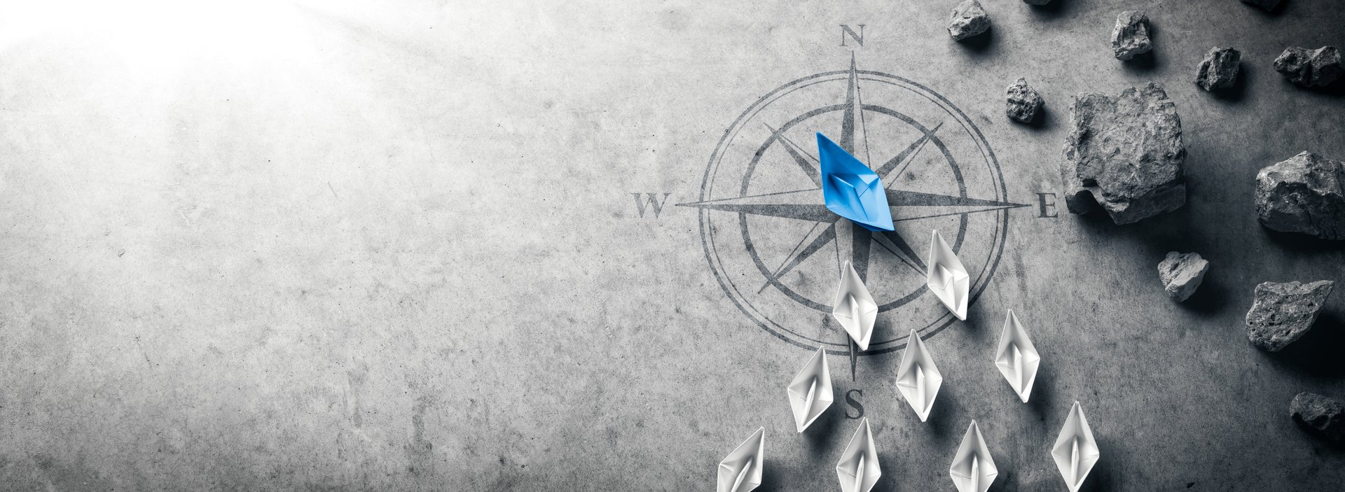 Blue Paper Boat With Compass Rose Leading A Fleet Of Small White Boats Away From Rocks On Grey Concrete - Leadership Concept