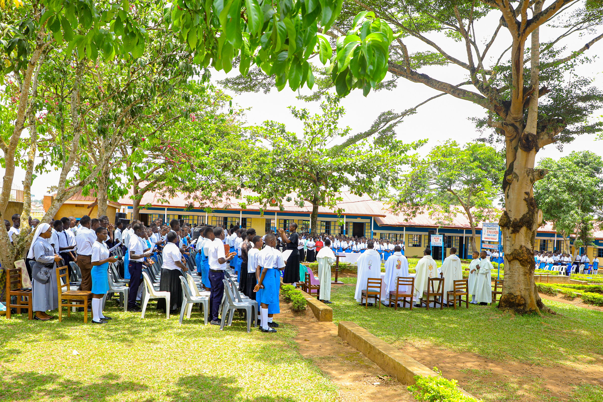 A group of students in uniforms attending an outdoor ceremony with priests under large trees on school grounds.