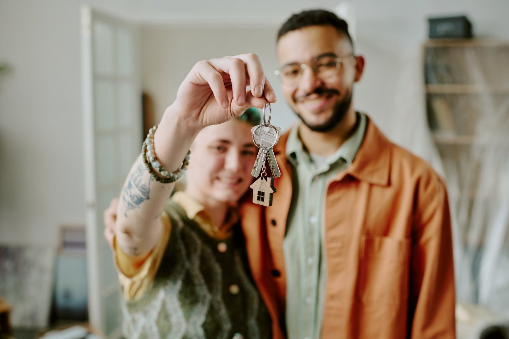 Young adult woman and man smiling and holding house keys together in home interior, showing keys toward camera, couple moving and doing renovations